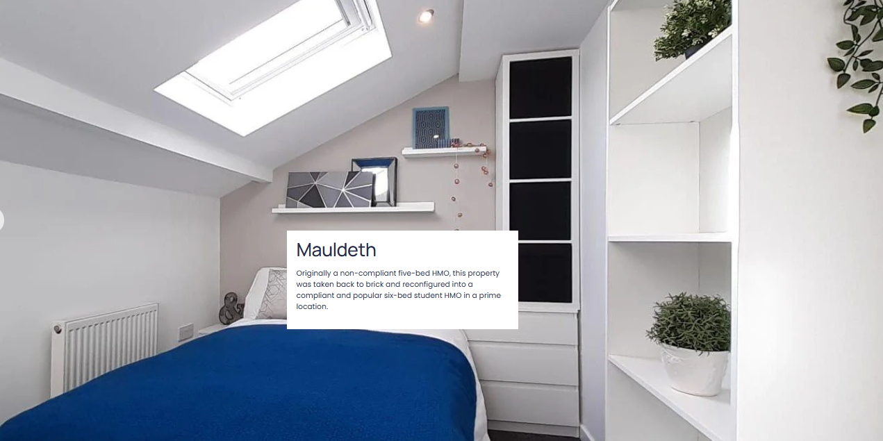 A cozy bedroom with a sloped ceiling and a skylight window. There is a bed with a blue blanket, a unique geometric mirror, and white shelves with decorative items and a potted plant.