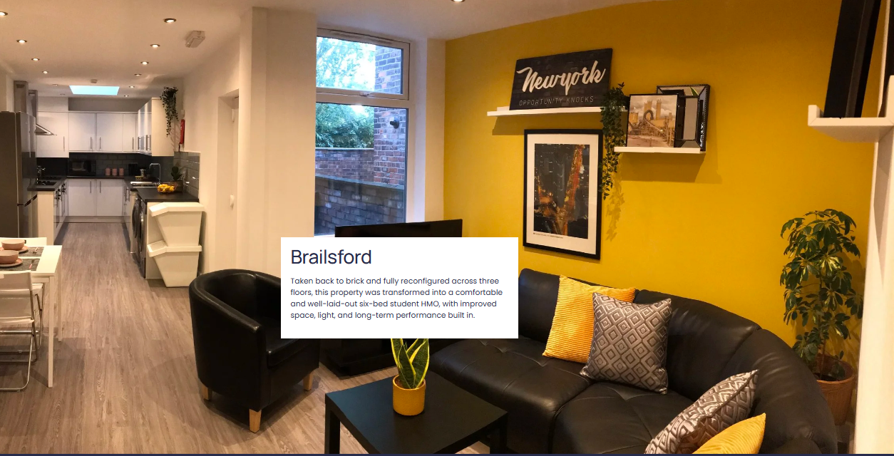 Living room with yellow accent wall, black leather sofa with patterned and yellow pillows, black armchair, small black coffee table with potted plant, framed artwork, and shelves with decorative items. Adjacent open kitchen with white cabinets and appliances.