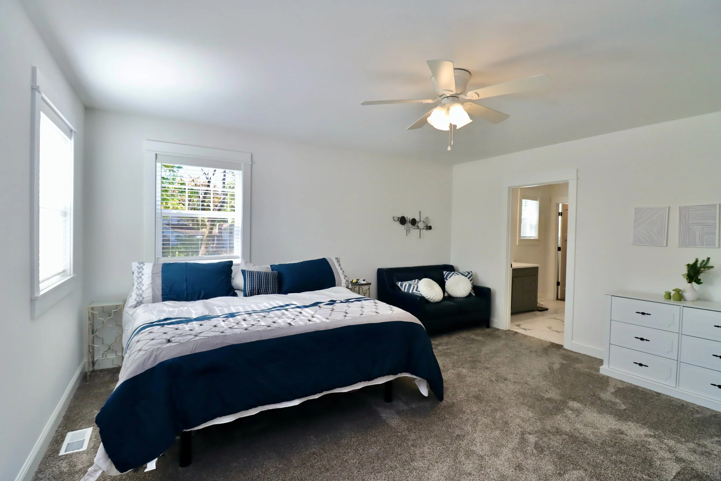 Bedroom with a bed that has blue and white bedding, two windows with white blinds, a ceiling fan with light, a small black sofa with white and blue pillows, a white dresser with green decor, and a doorway leading to an adjacent room.