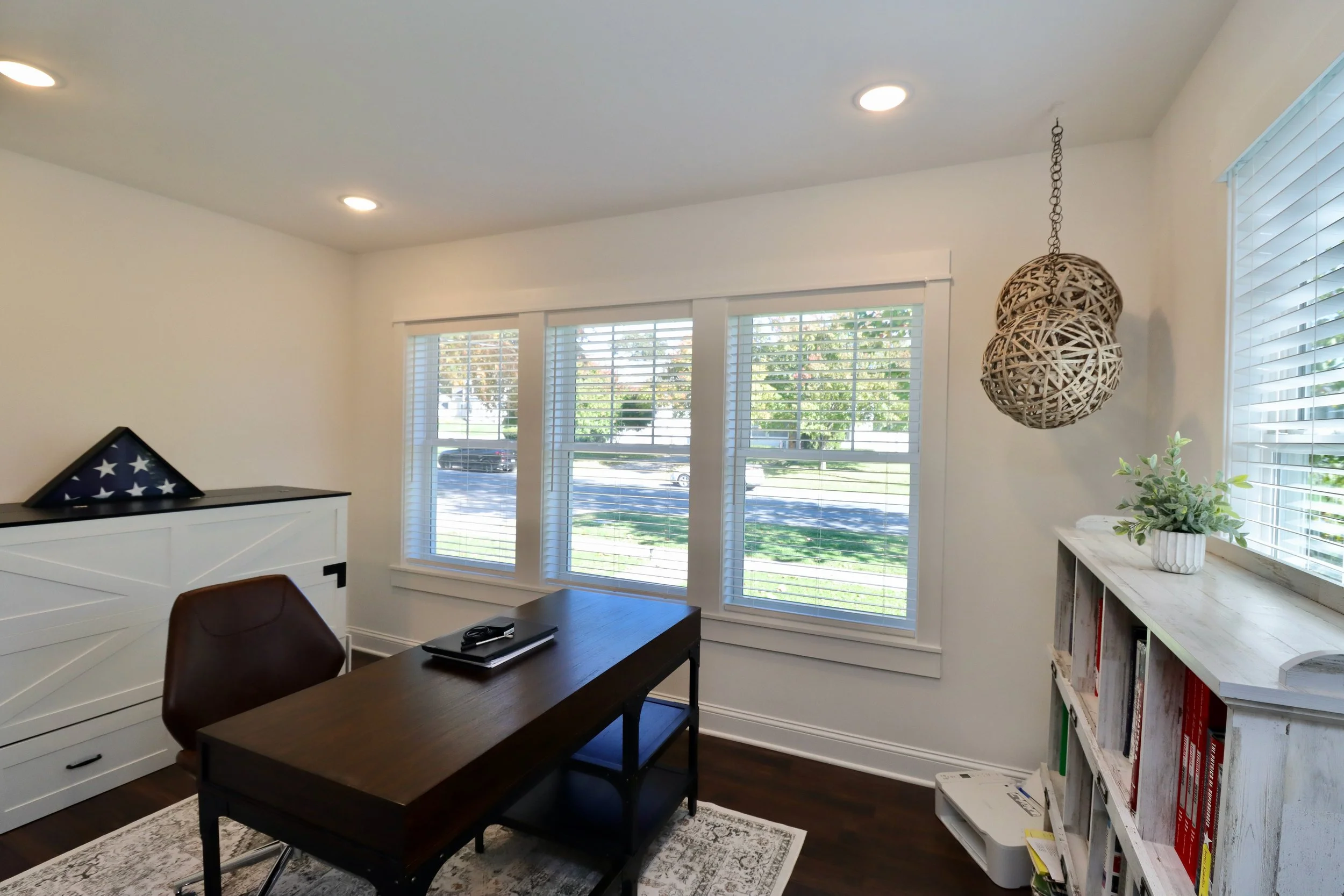 Home office with large windows, white walls, dark wood desk, brown chair, white bookshelf, decorative hanging wicker lamps, and potted plant.