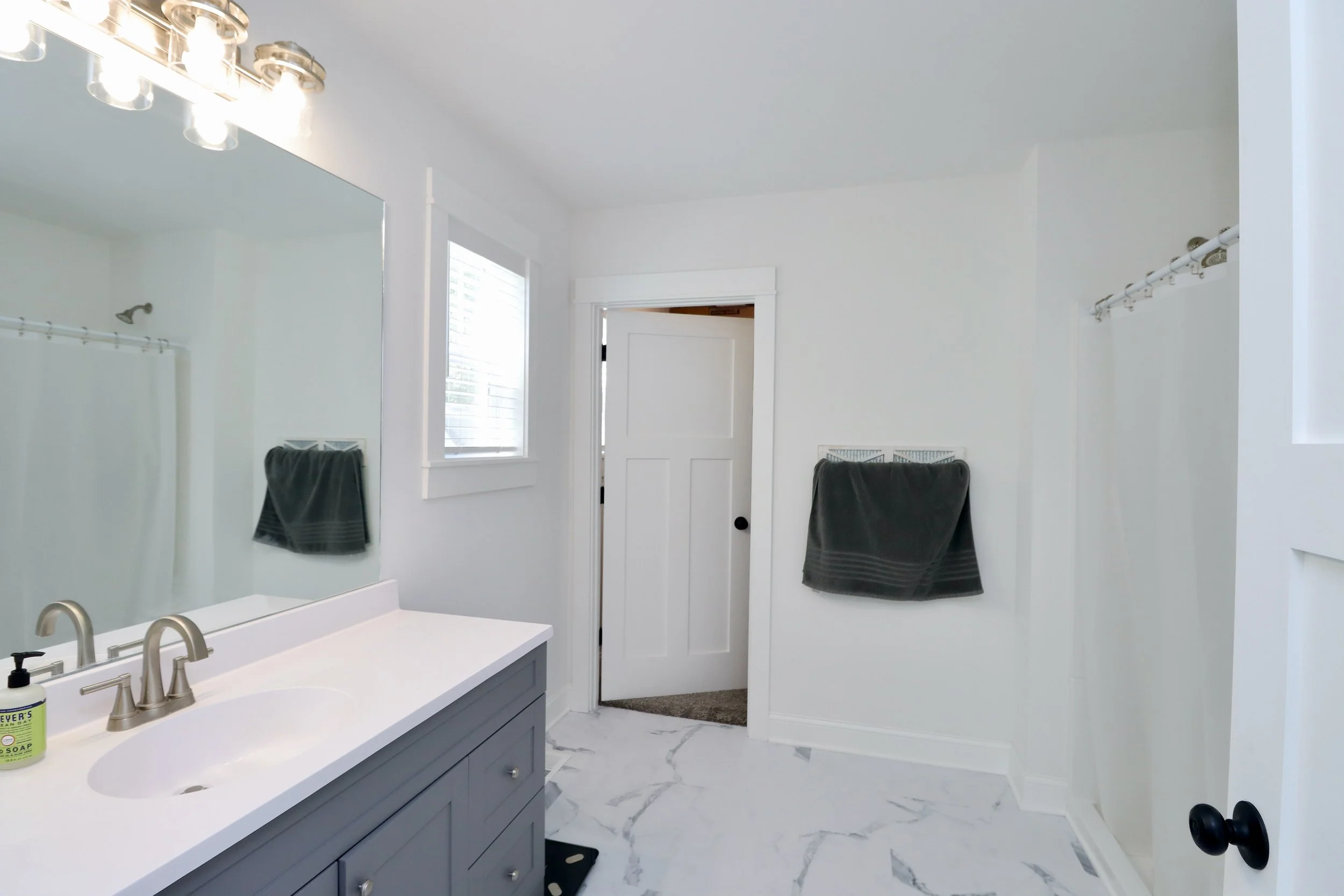 A bathroom with a white wall and marble floor, gray vanity with a white countertop, a large mirror, a window with blinds, a doorway slightly open, gray towels on racks, and a shower area with a curtain rod.