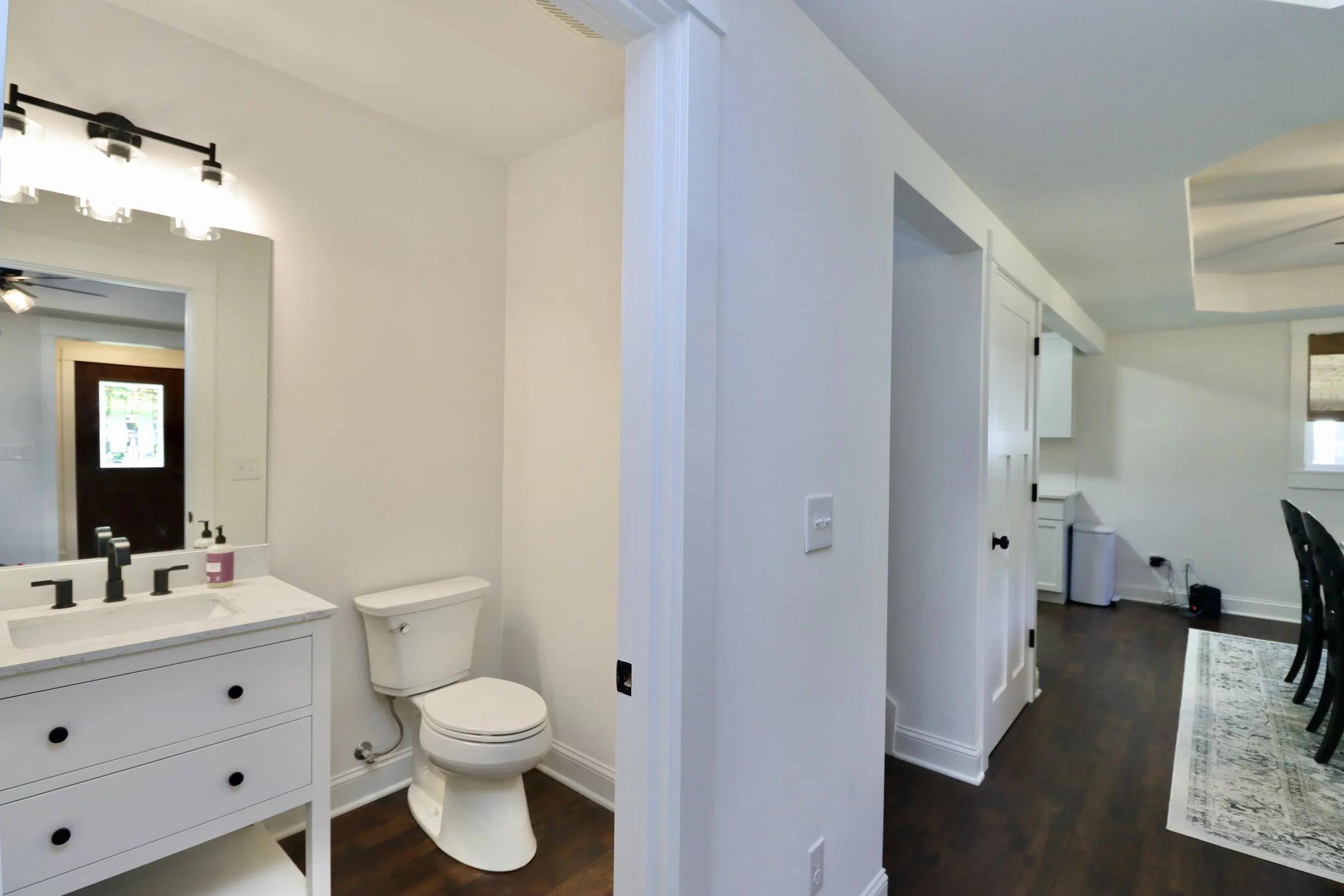 Bathroom with white vanity, black fixtures, behind mirror, and toilet on white wall; adjacent to a dining area with dark wood floors, black chairs, and a patterned rug.