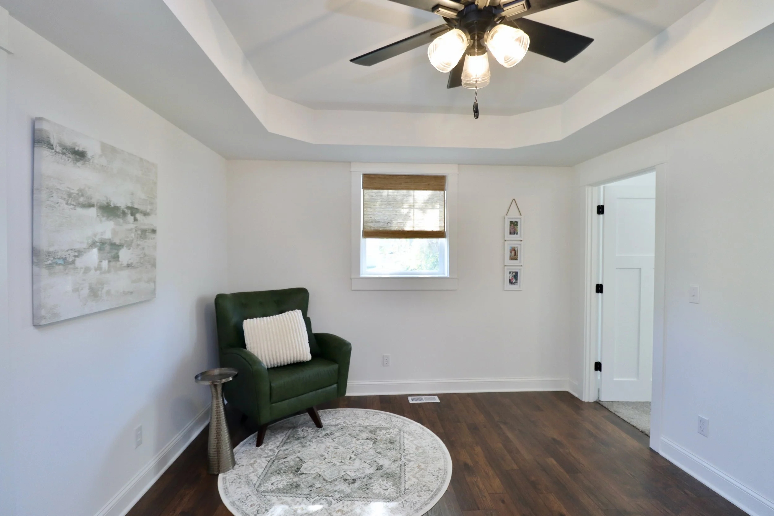 A living room with a green armchair, a white pillow, a silver side table, a round patterned rug, a window with a brown blind, a white wall with framed pictures, a ceiling fan with lights, and hardwood flooring.