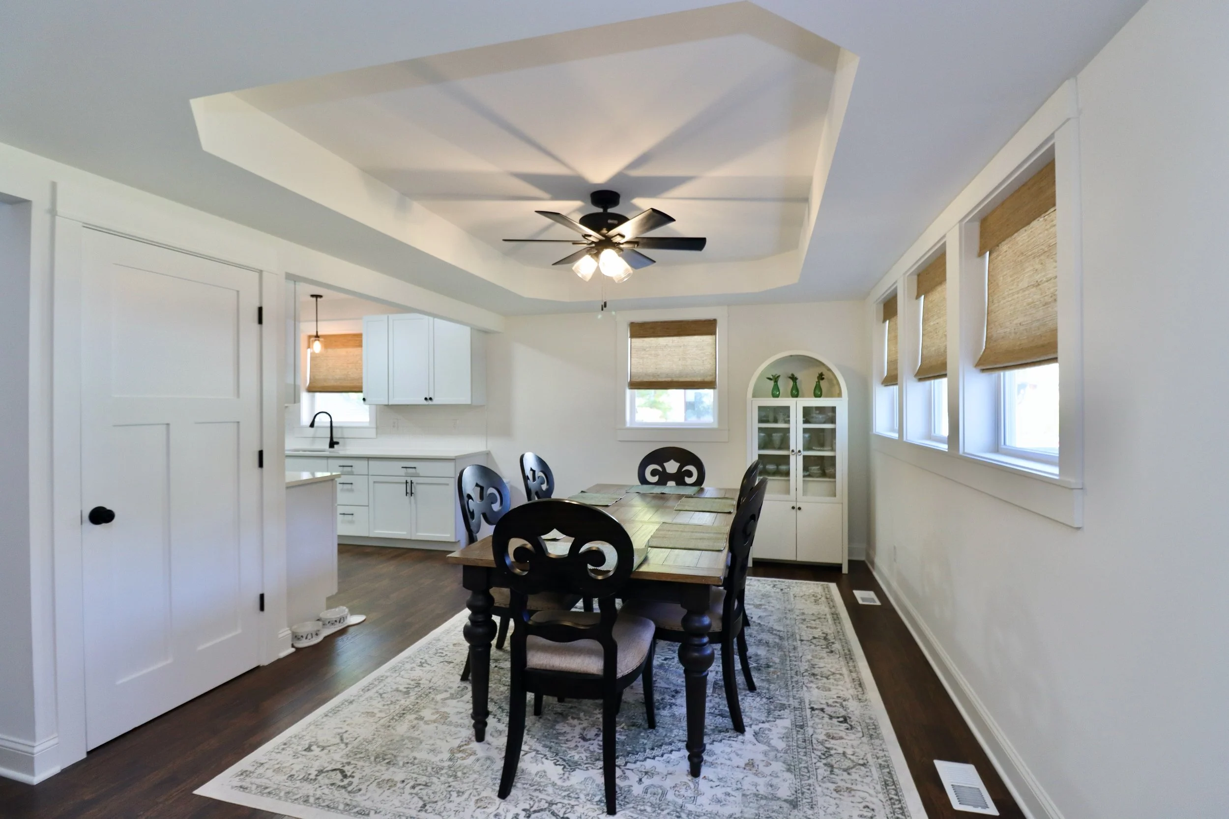 Dining room with a dark wood table, six matching chairs with decorative backs, a white cabinet with glass doors, and three windows with beige blinds. The room has a white ceiling with a black ceiling fan and light, and a patterned area rug on dark wo