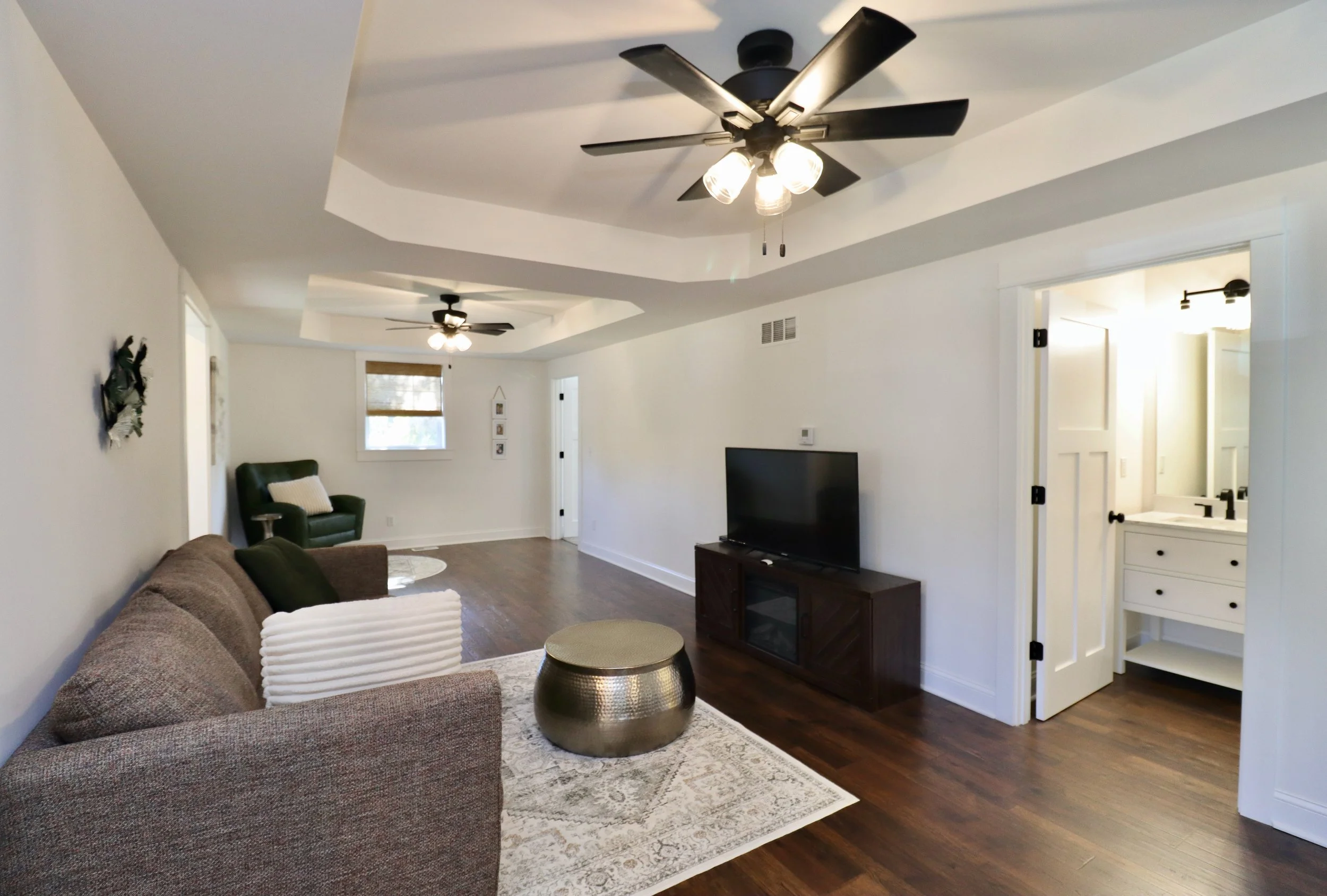 Living room with white walls, hardwood floors, a brown couch with green pillows, a brass-colored round coffee table, a patterned area rug, a TV on a wooden stand, and a small dark green armchair by a window with a partial shade. There are two ceiling