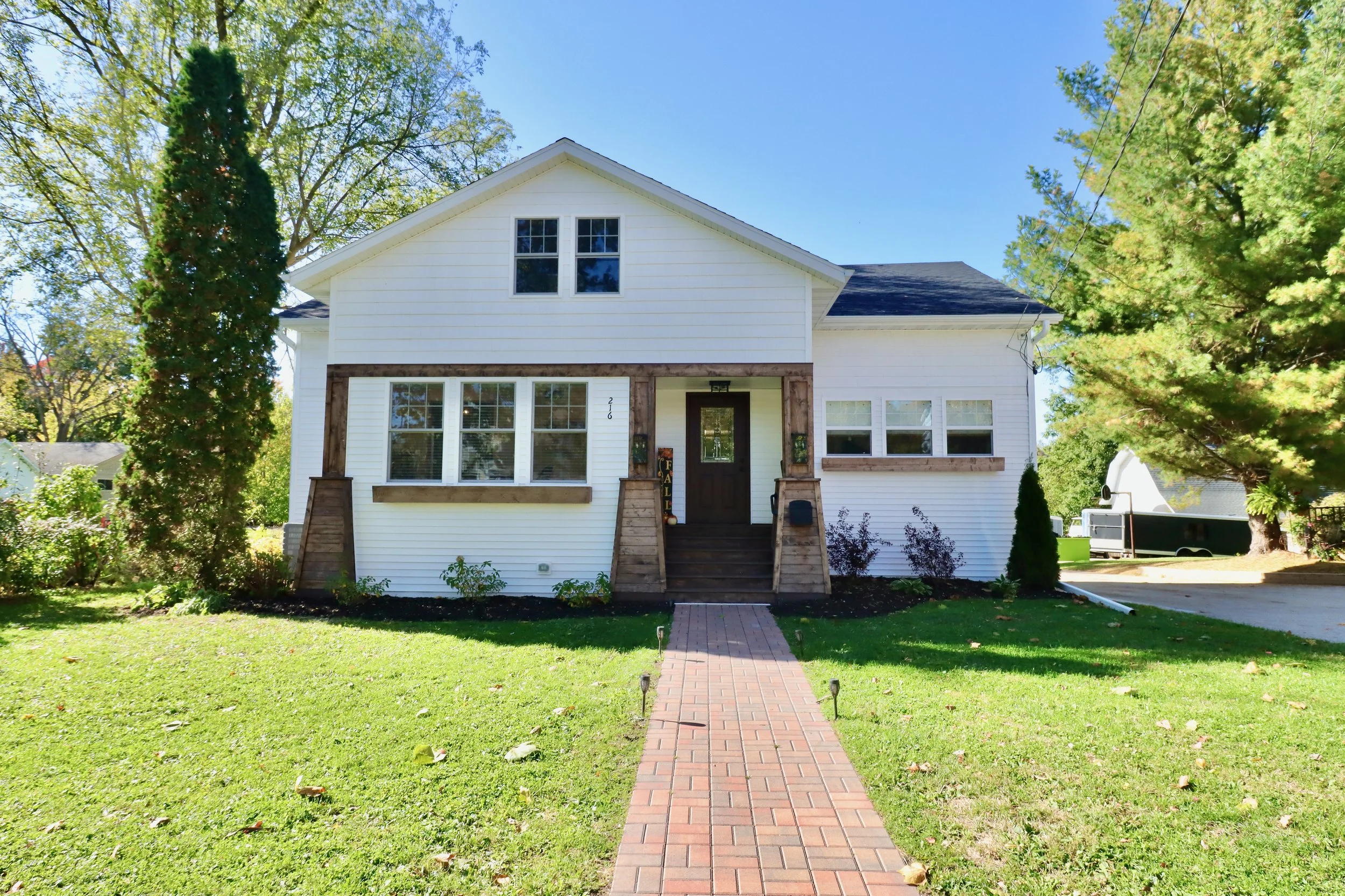Front view of a two-story white house with a brick walkway leading to the front door, surrounded by green grass, trees, and shrubs, on a sunny day.