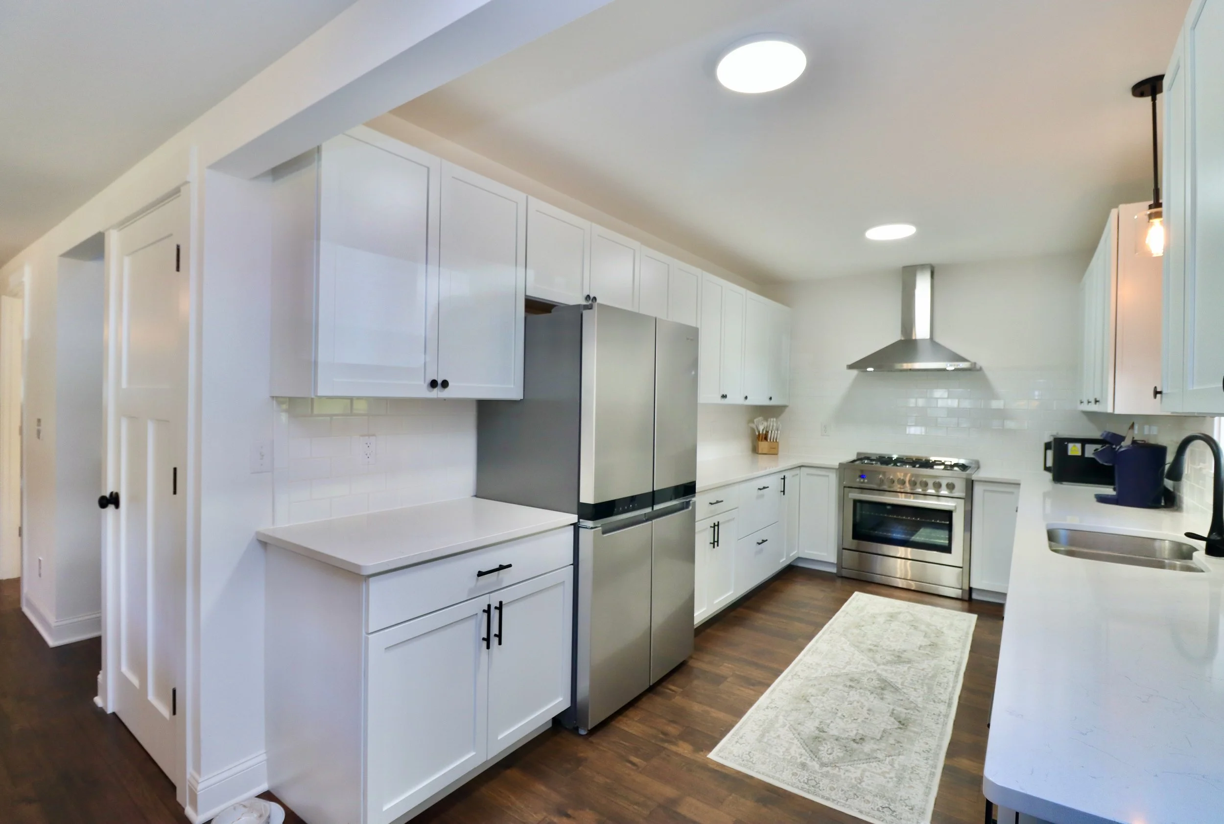 Modern kitchen with white cabinets, stainless steel refrigerator and oven, dark wood flooring, white backsplash, and a light-colored rug on the floor.