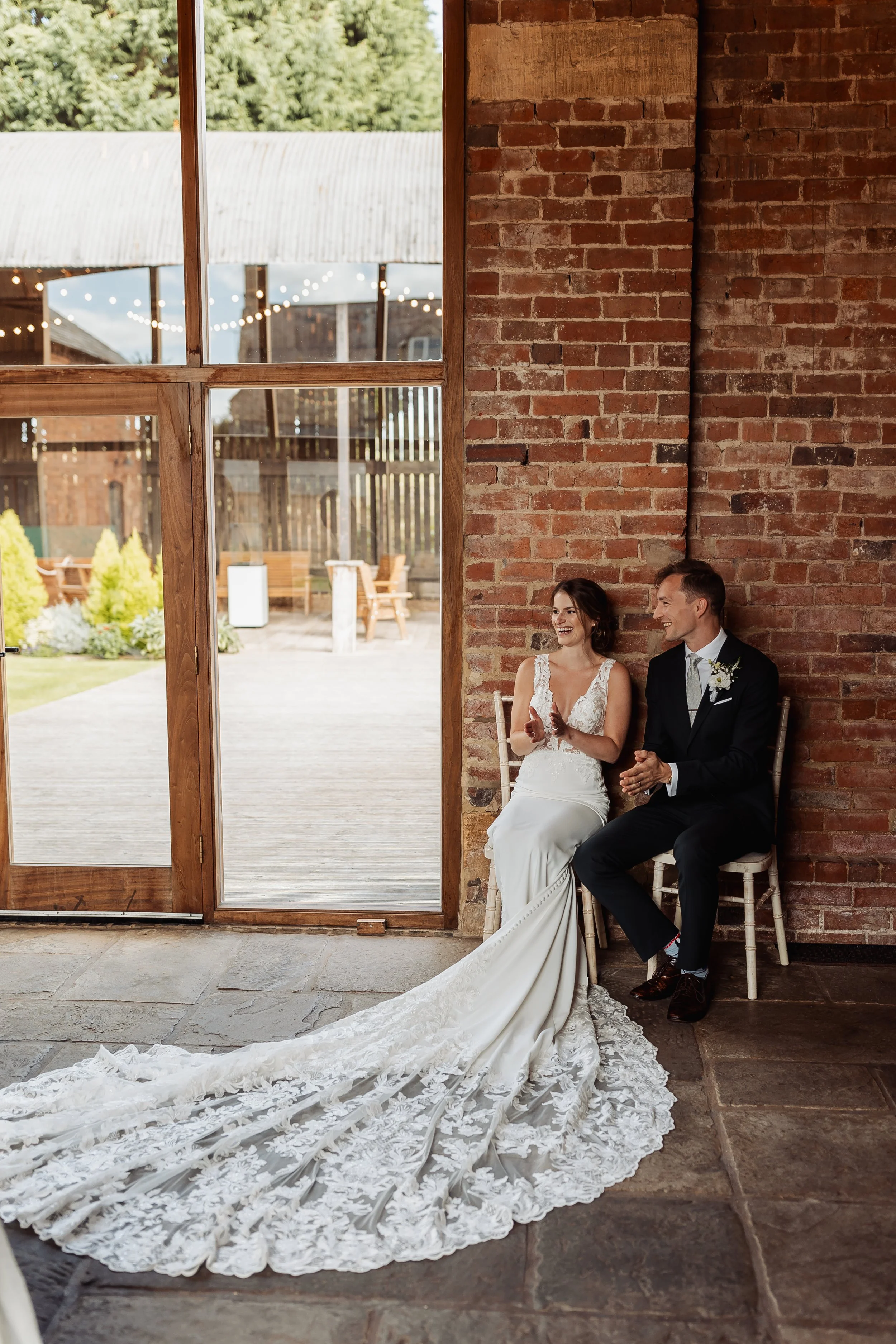 A newlywed couple sitting on chairs inside a rustic venue with a brick wall and large window, celebrating after their wedding.