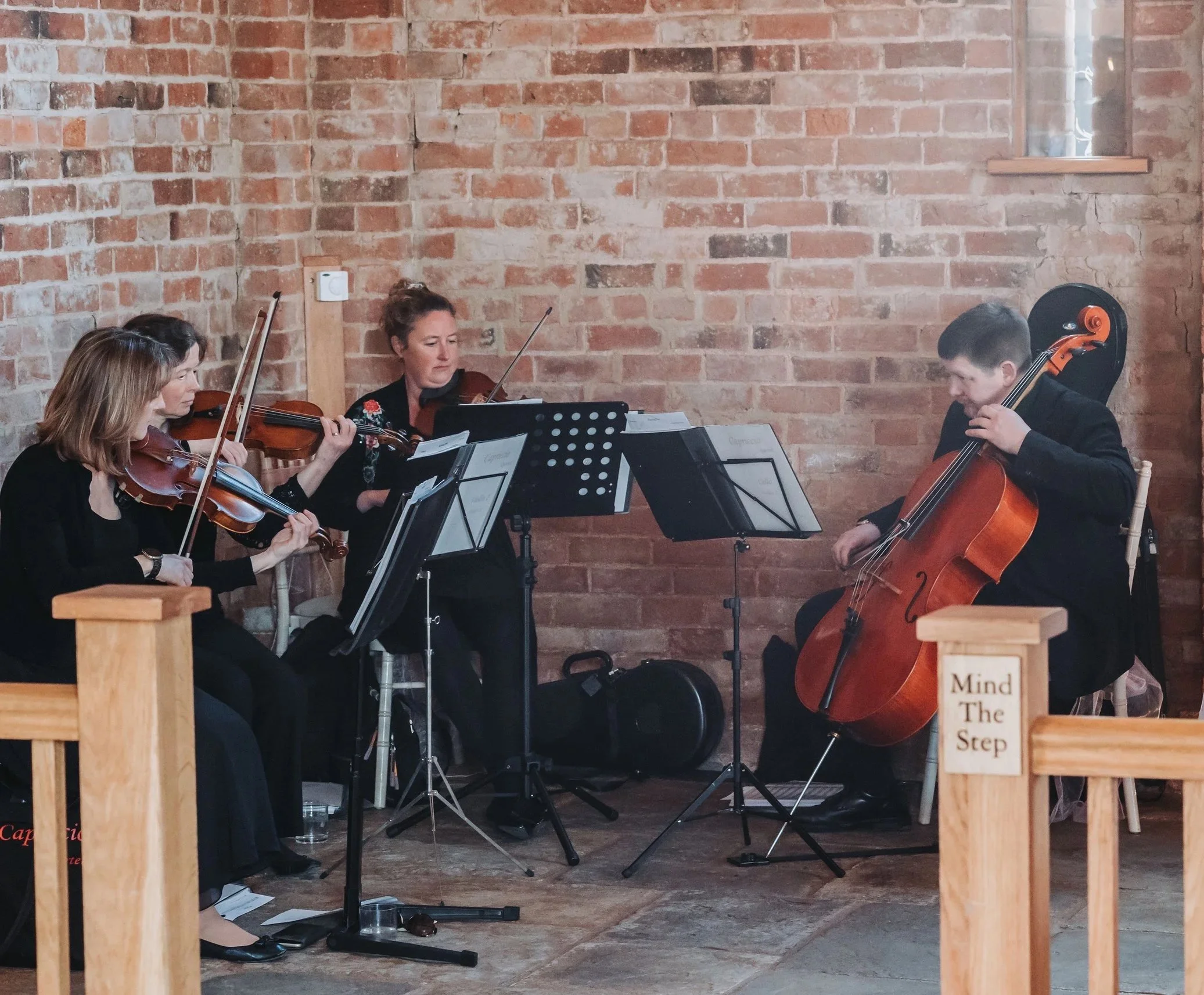 Three women and one man sitting and playing classical string instruments in a room with exposed brick walls. The women are playing violins and the man is playing a cello. They have music stands in front of them.