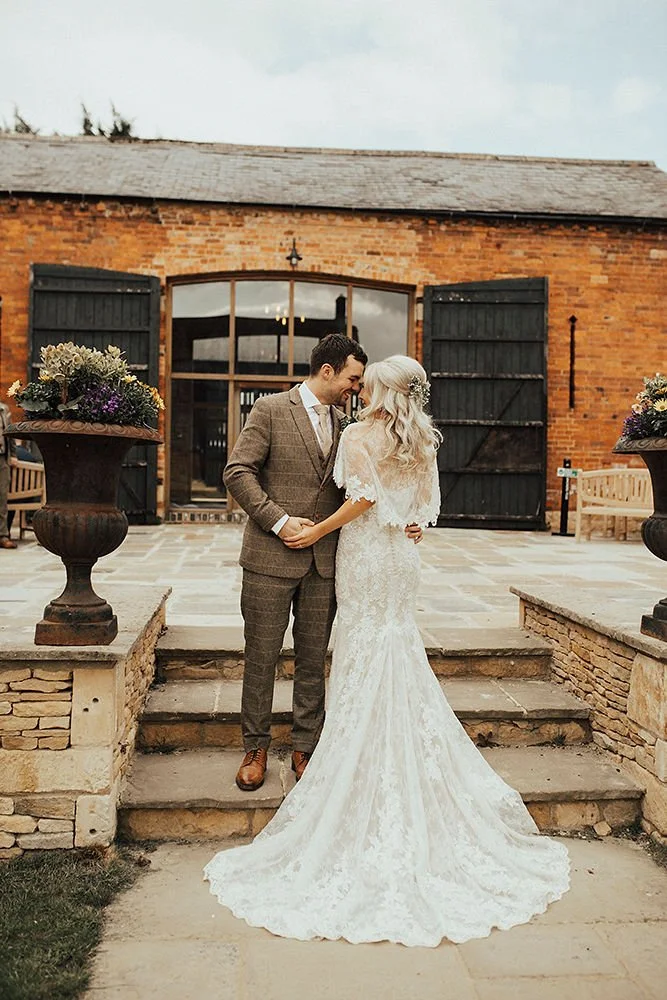 Bride and groom standing on steps outside a rustic building, smiling and holding hands, with early evening lighting.