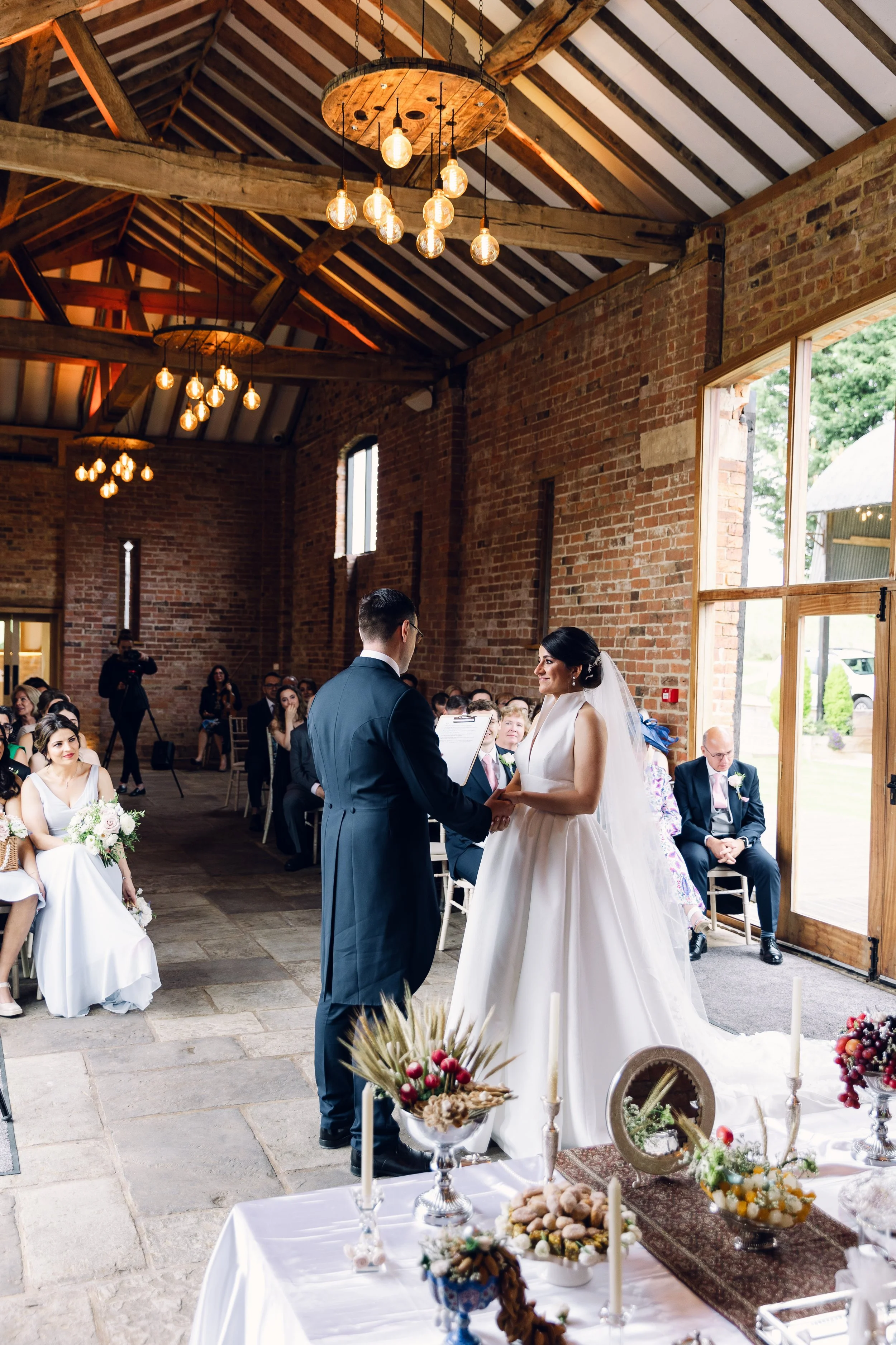 Bride and groom exchanging vows during wedding ceremony in rustic indoor venue with brick walls, wooden beams, and hanging light fixtures, while guests look on.