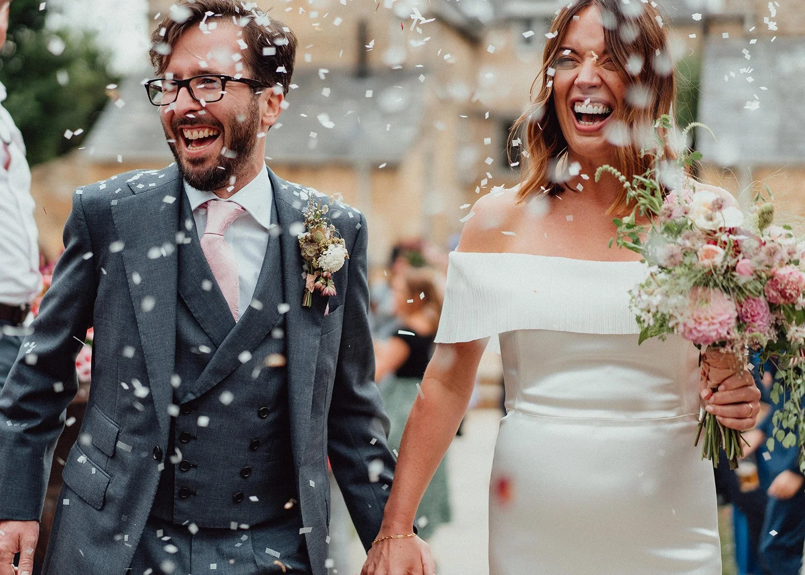 A joyful bride and groom at their wedding celebration, holding hands and surrounded by falling confetti.