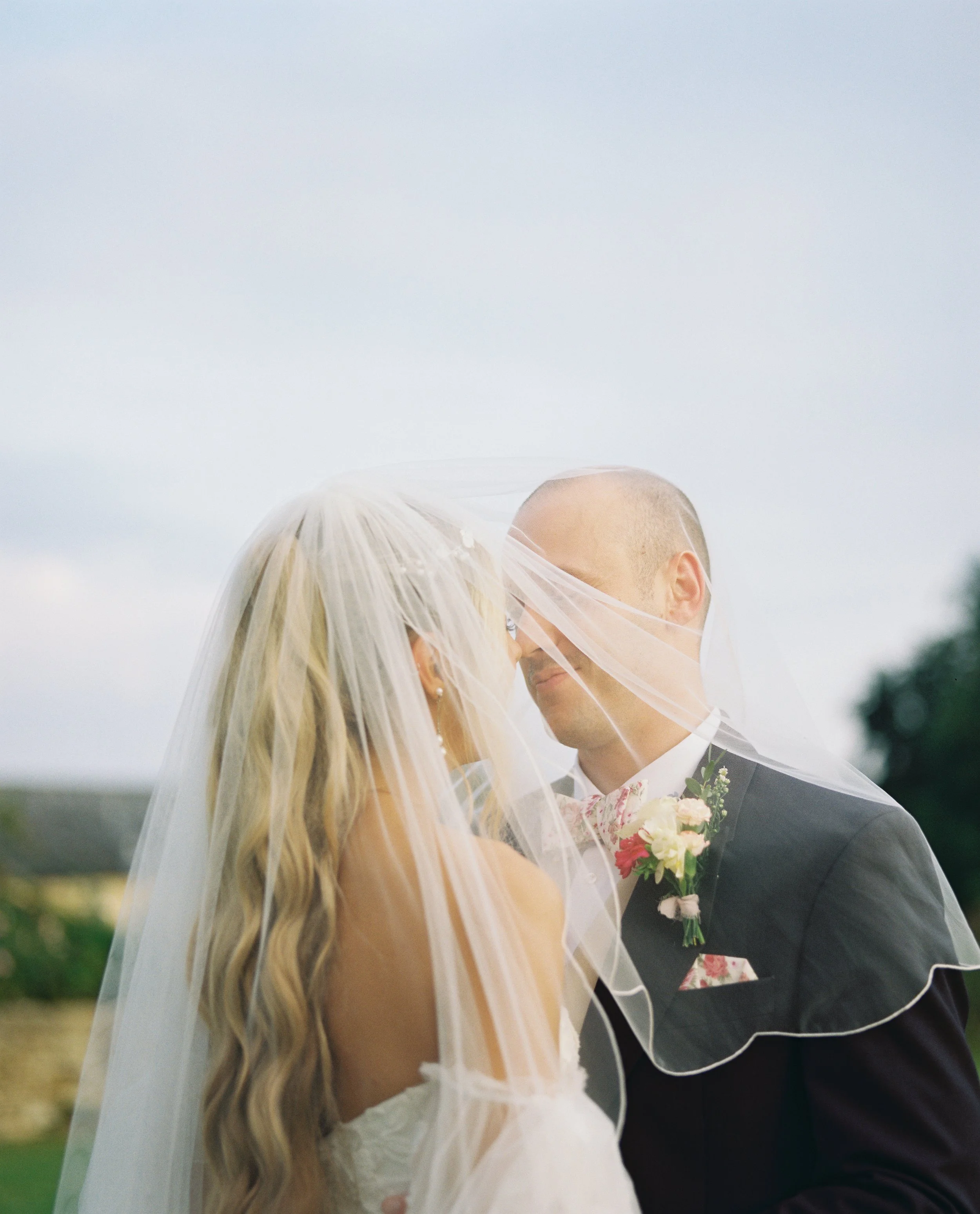 A bride and groom embrace under a veil on their wedding day outdoors.