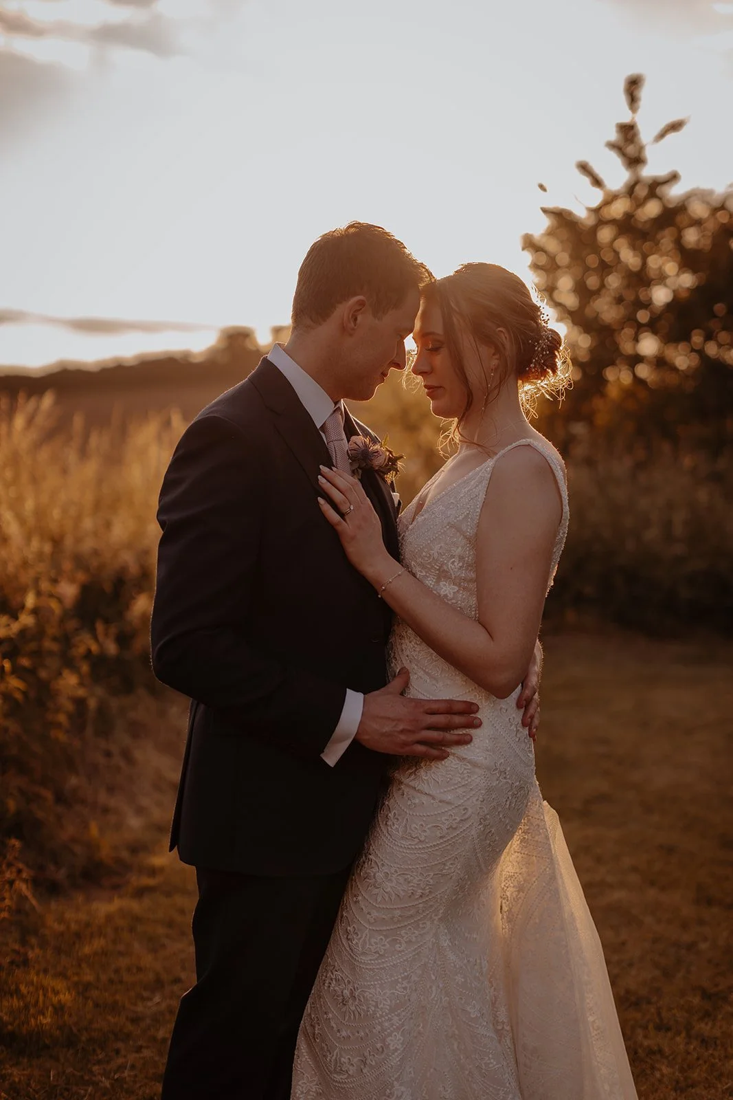 A bride and groom embracing each other outdoors at sunset, with head touching, in wedding attire. The bride is wearing a lace wedding dress, the groom a dark suit with a boutonniere. The background features a field and trees illuminated by warm golden light.