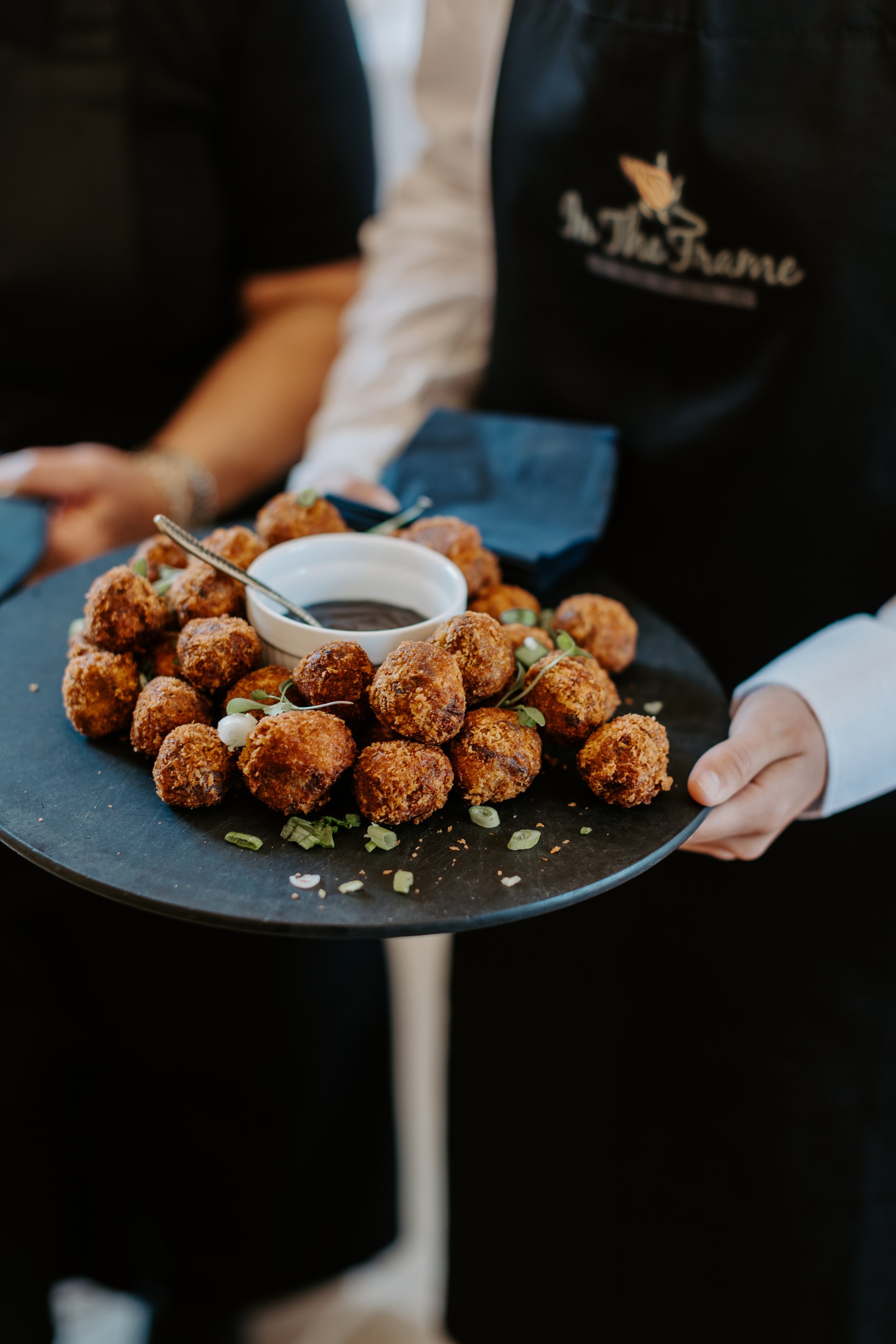 A server holding a black tray with fried appetizers, possibly chicken nuggets or croquettes, garnished with green onions and a small bowl of dipping sauce.