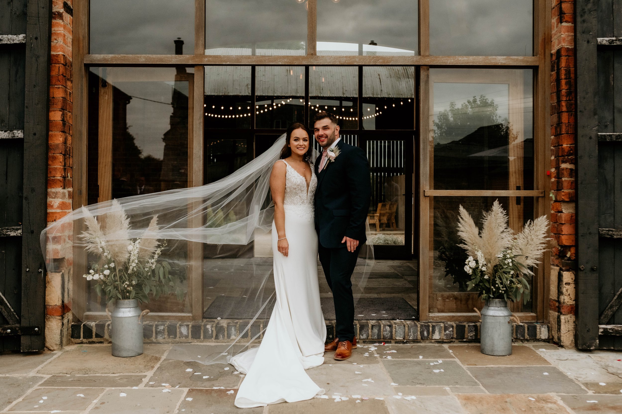 Bride and groom standing together in front of a rustic glass door with string lights inside. The bride is wearing a white wedding gown and veil, and the groom is dressed in a dark suit with a tie. Two vases with pampas grass and white flowers are on either side of the entrance. Petals are scattered on the ground.