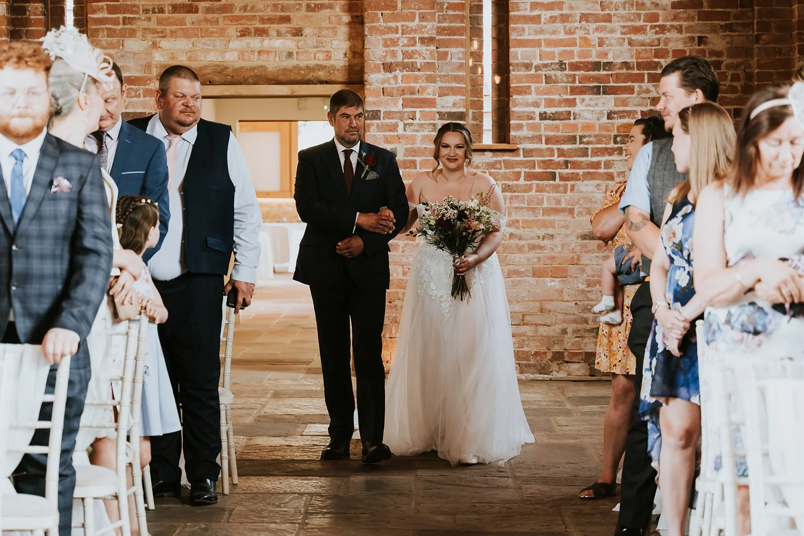A bride walking down the aisle with her father at a wedding in a rustic venue with brick walls, holding a bouquet of flowers, with guests seated on either side watching.