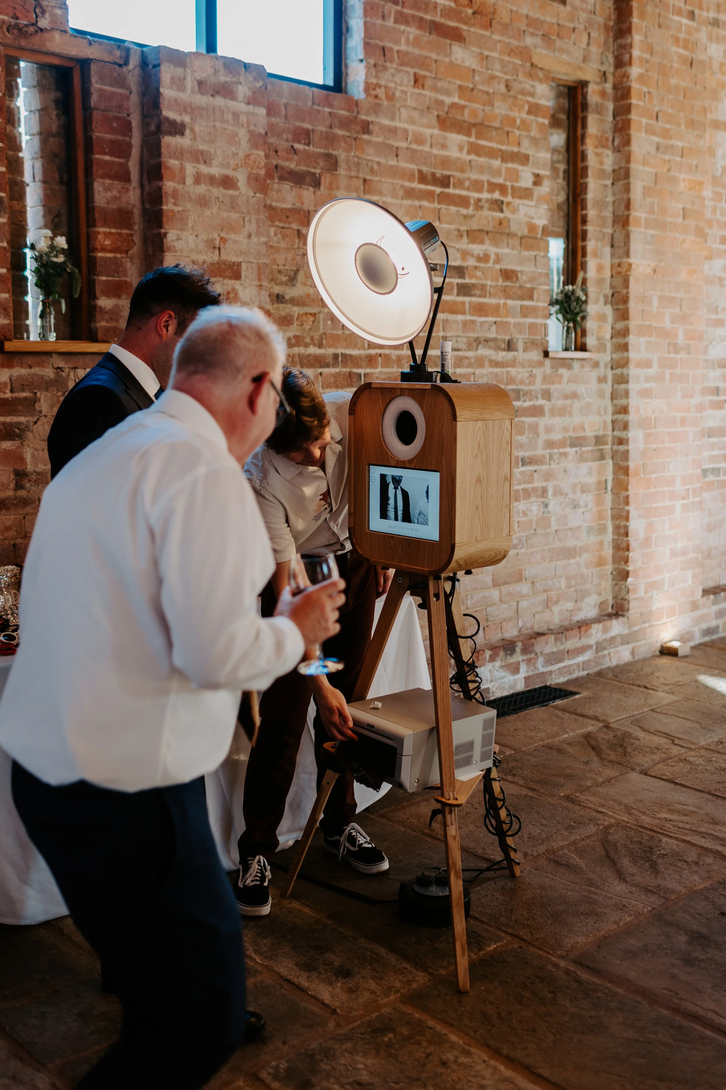 People at a photo booth during a celebration in a rustic indoor setting with brick walls and small windows.