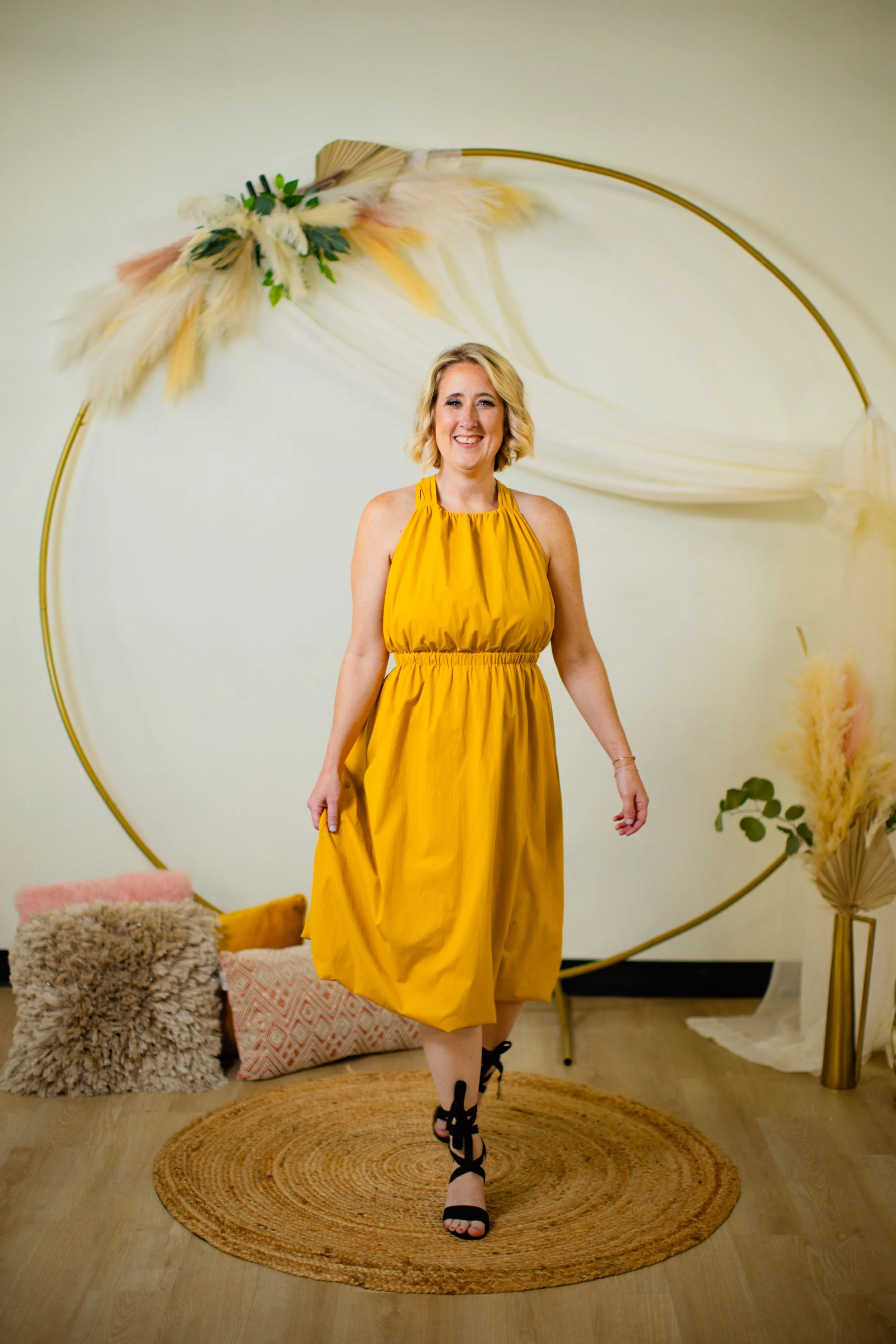A woman smiling and walking in front of a decorative backdrop with a large gold circle, fabric, and feather decorations. She is wearing a yellow dress and black high-heeled lace-up sandals.