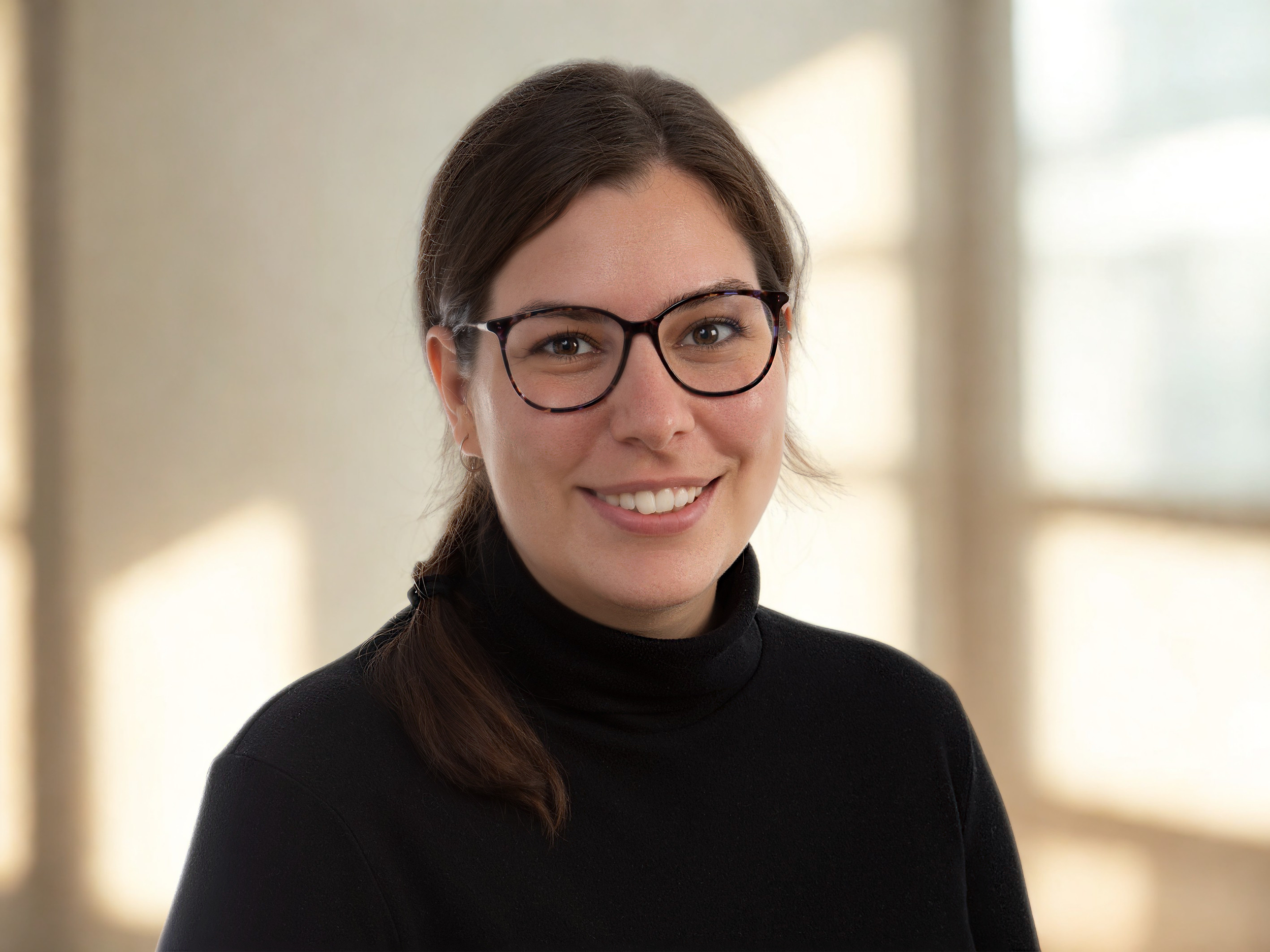 A woman with brown hair, glasses, and a black turtleneck sweater smiling at the camera, standing in a softly lit indoor setting.