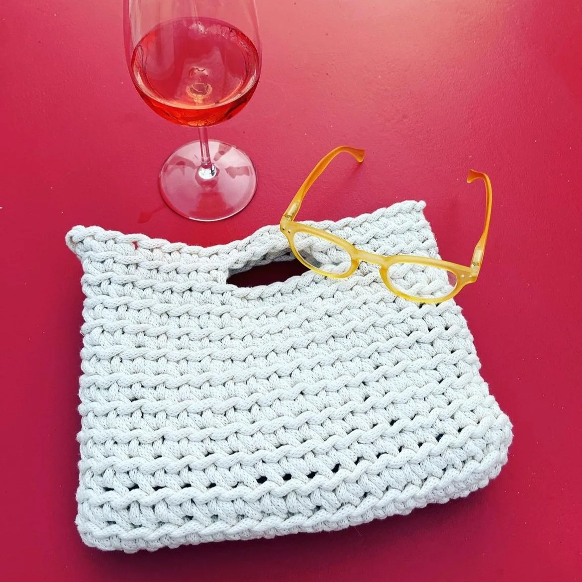 Ein Glas mit Roséwein, eine weiße gestrickte Tasche, eine gelbe Sonnenbrille, auf rotem Hintergrund.