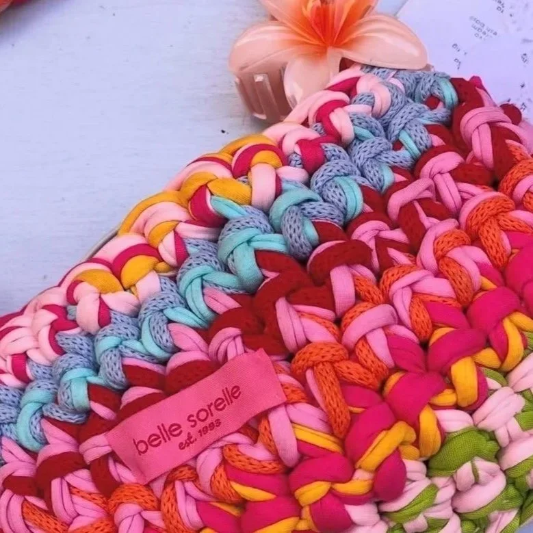 Bunte, handgemachte Deko-Kette aus geflochtenen Stoffstreifen mit einem Belle Sorelle Etikett, auf einem weißen Oberflächen, umgeben von Blumen und Papier.