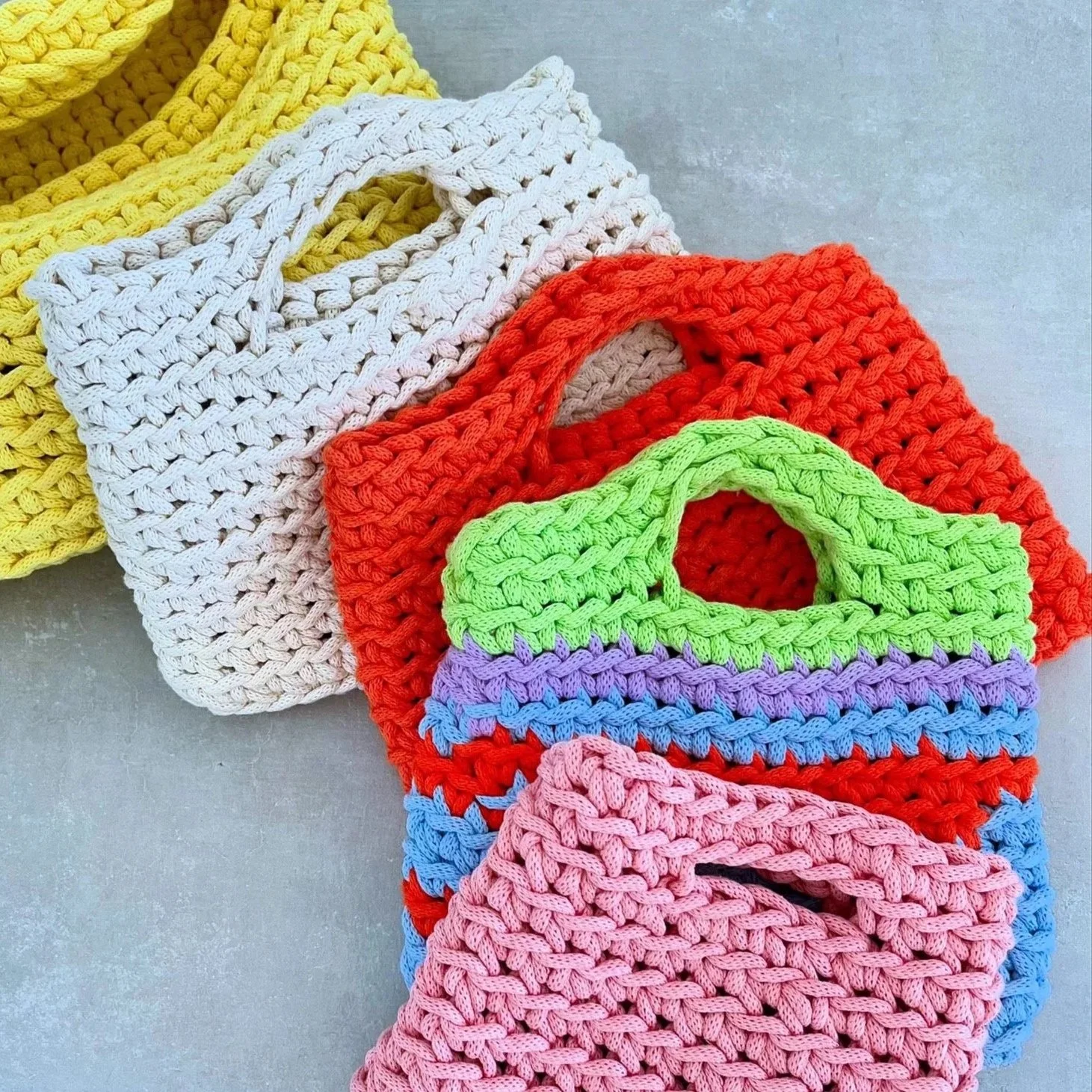 Bunte, handgefertigte gestrickte Taschen in verschiedenen Farben, gelb, weiß, rot, grün, lila, blau, rosa, auf einer hellgrauen Oberfläche.