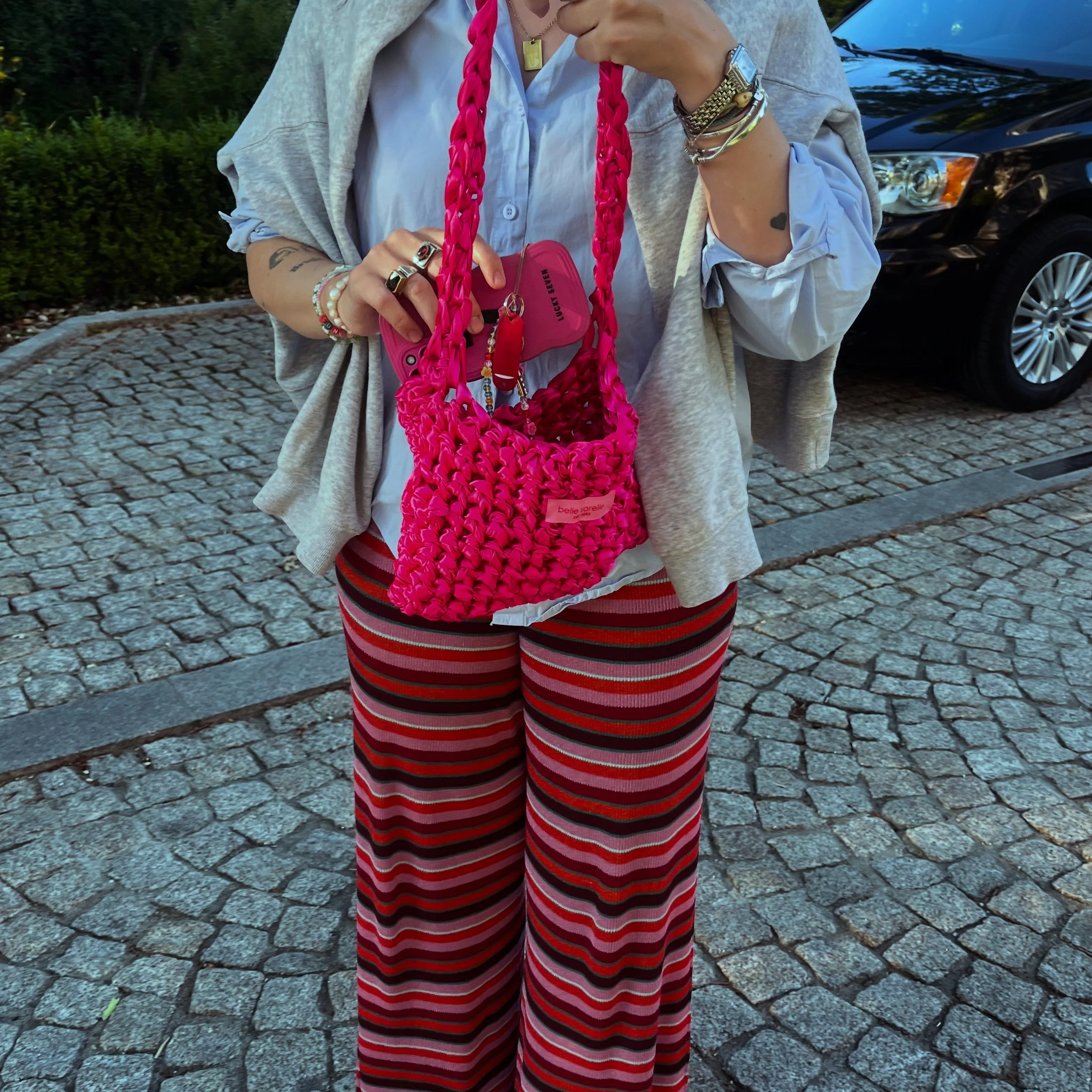 Eine Person trägt eine weiße Hemd- und eine graue Jacke, hat mehrere Schmuckstücke und Ringe an den Händen, hält eine pinkfarbene, gestrickte Tasche mit einem Schlüsselbund darin, trägt rote, schwarze und rosa gestreifte weite Hosen, steht auf einem gepflasterten Boden.