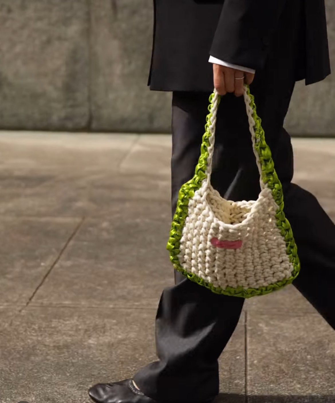 Person trägt eine handgemachte Tasche aus geflochtenem, dickem Garn mit grünen und weißen Farbdetails, auf einem Bürgersteig bei Nacht.