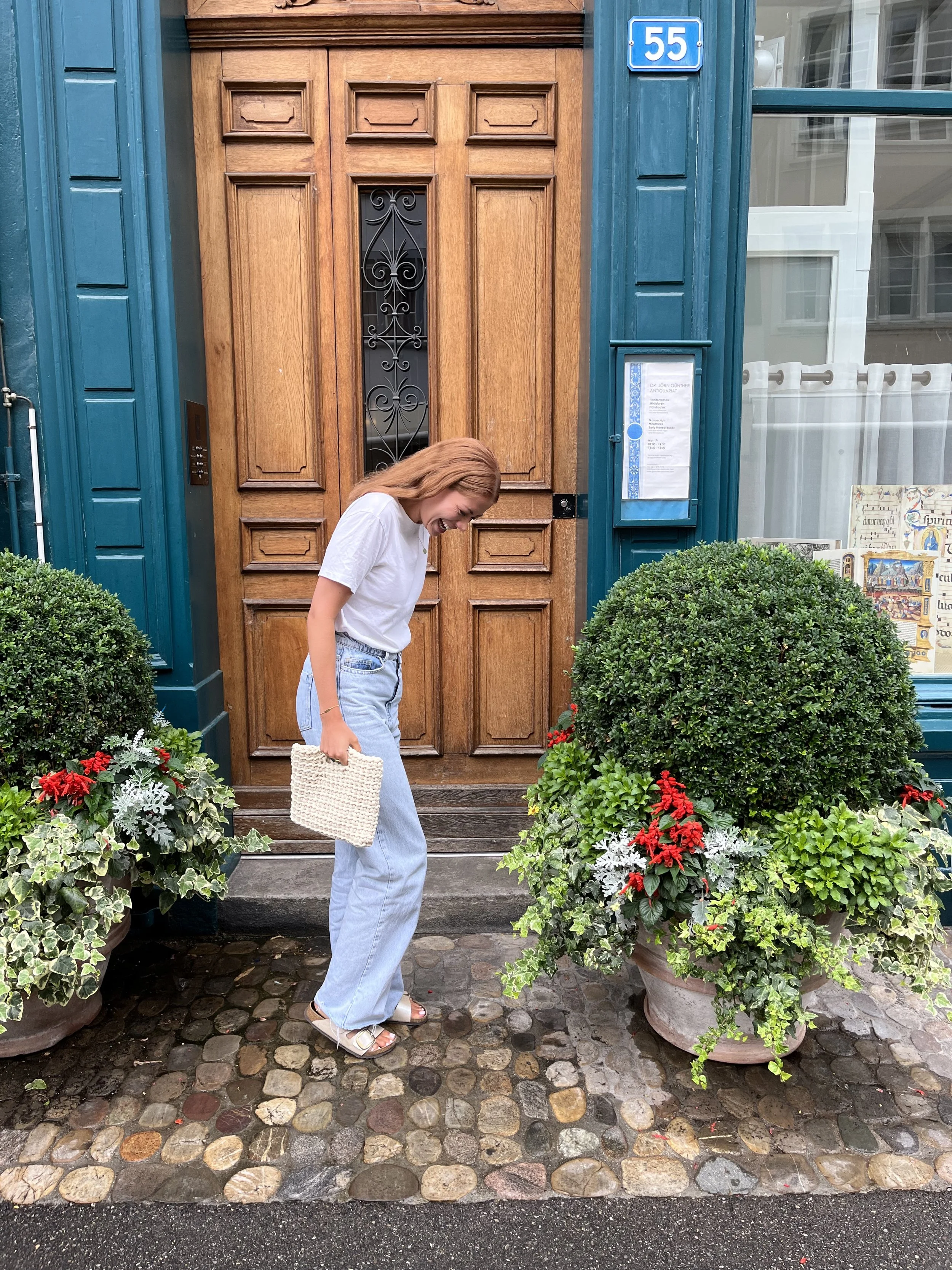 Junge Frau mit lockigem, rotem Haar trägt weiße T-Shirts, helle Jeans, Sandalen und einen weißen Korb auf dem Weg zum Eingang eines Hauses mit großen Topfpflanzen vor der Tür.