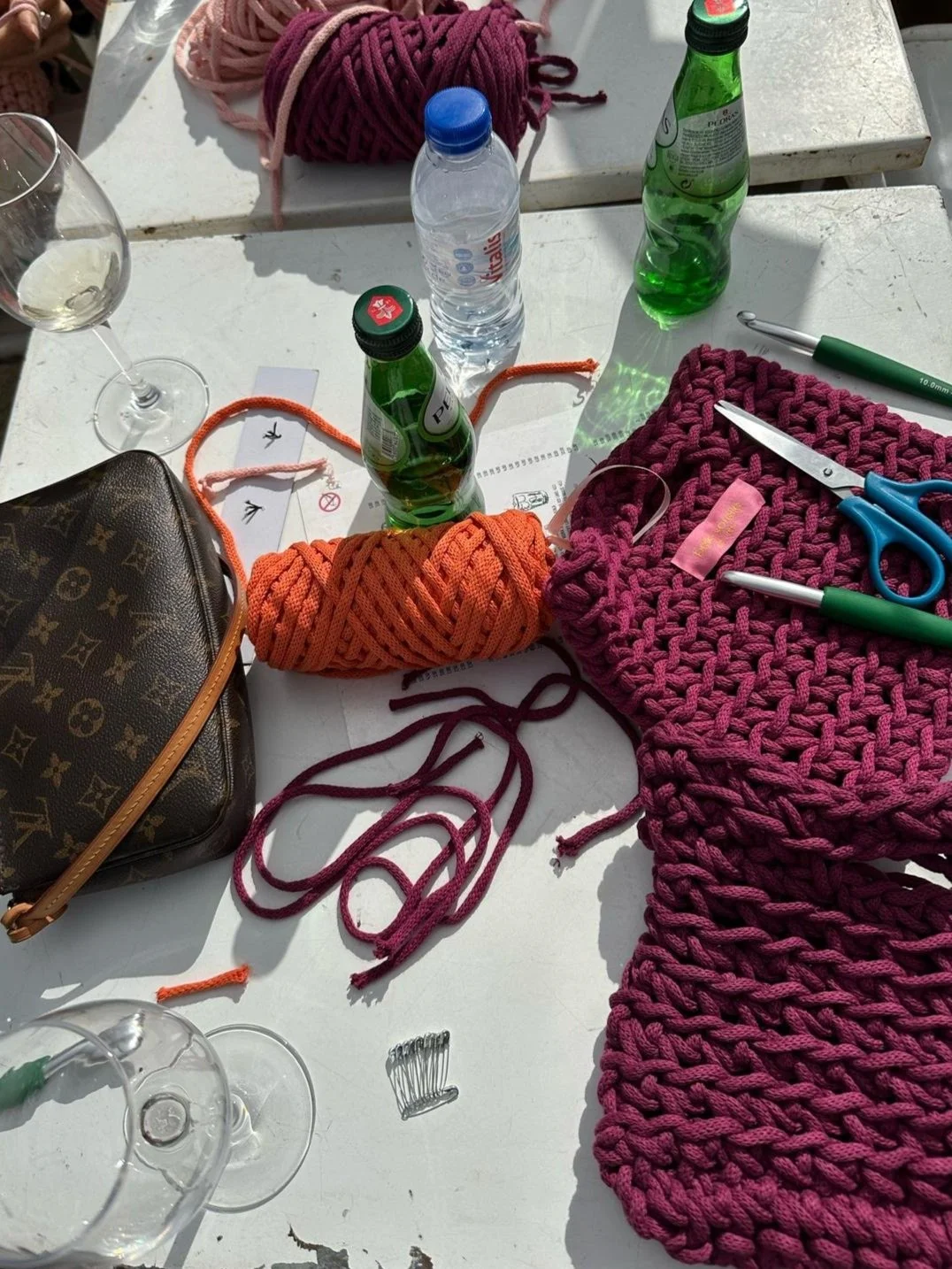 Strickwolle in Pink, Orange, und Rot, eine Schere, ein Messer, eine Flasche Wasser, eine Wein- und zwei Biergläser, eine Louis Vuitton Tasche, Schnittmuster, und Nähutensilien auf einem weißen Tisch.