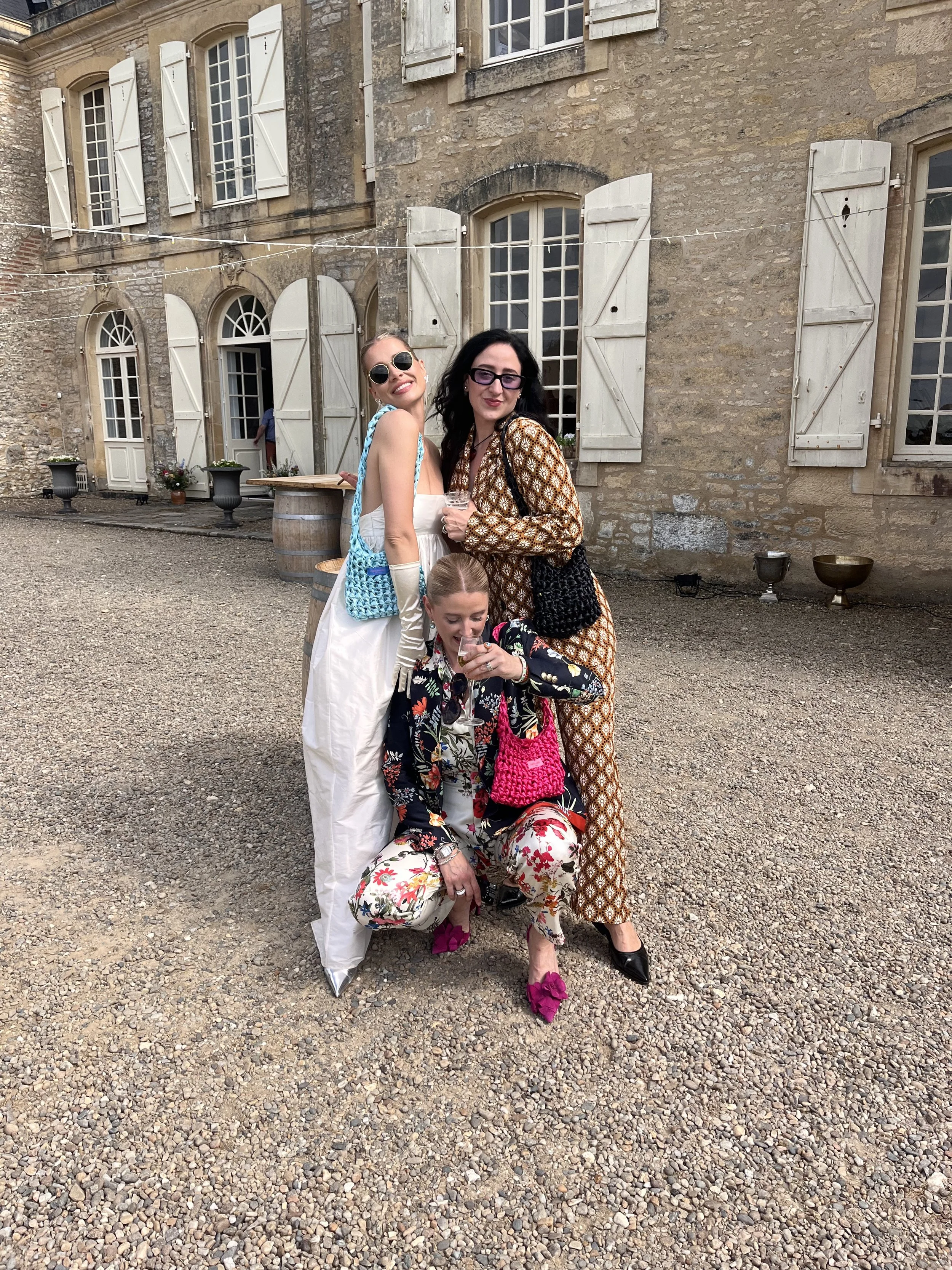 Drei Frauen posieren vor einem alten Stein-Gebäude mit weißen Fensterläden, während sie lachen und sich in bunten Kleidern und Accessoires präsentieren.