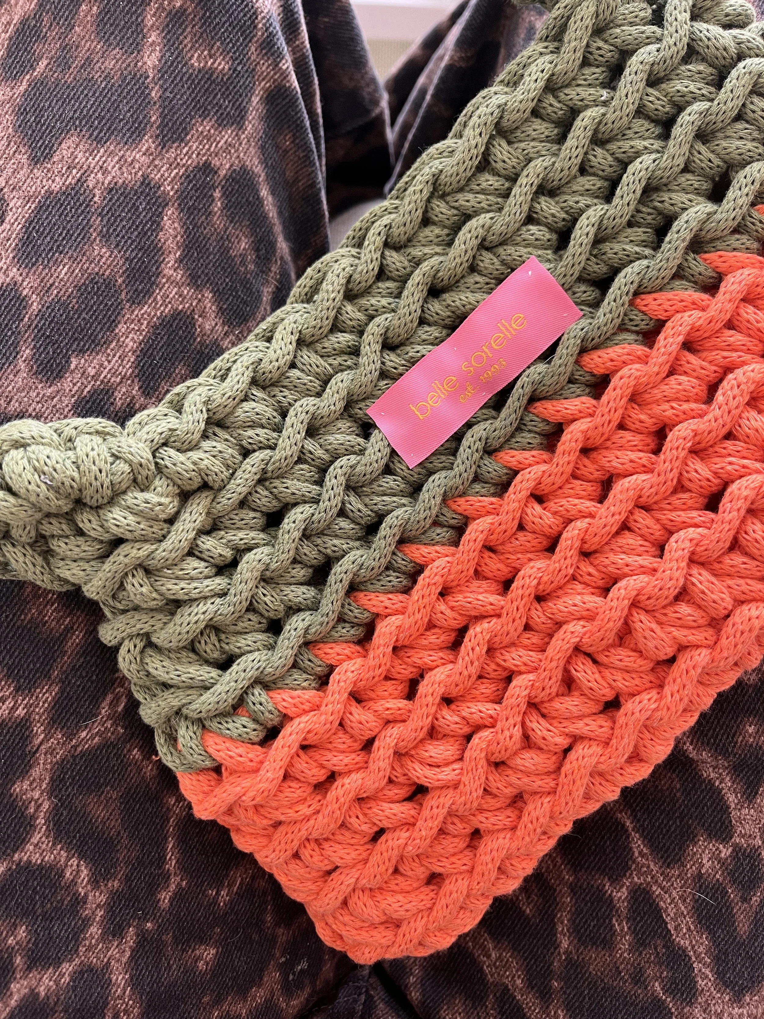 Nahaufnahme von zwei gestrickten Taschen, eine in olivgrün und eine in leuchtendem Orange, mit einem pinkfarbenen Etikett, das 'béli soréte seit 1992' trägt, auf einem dunklen Stoff mit Leopardenmuster.