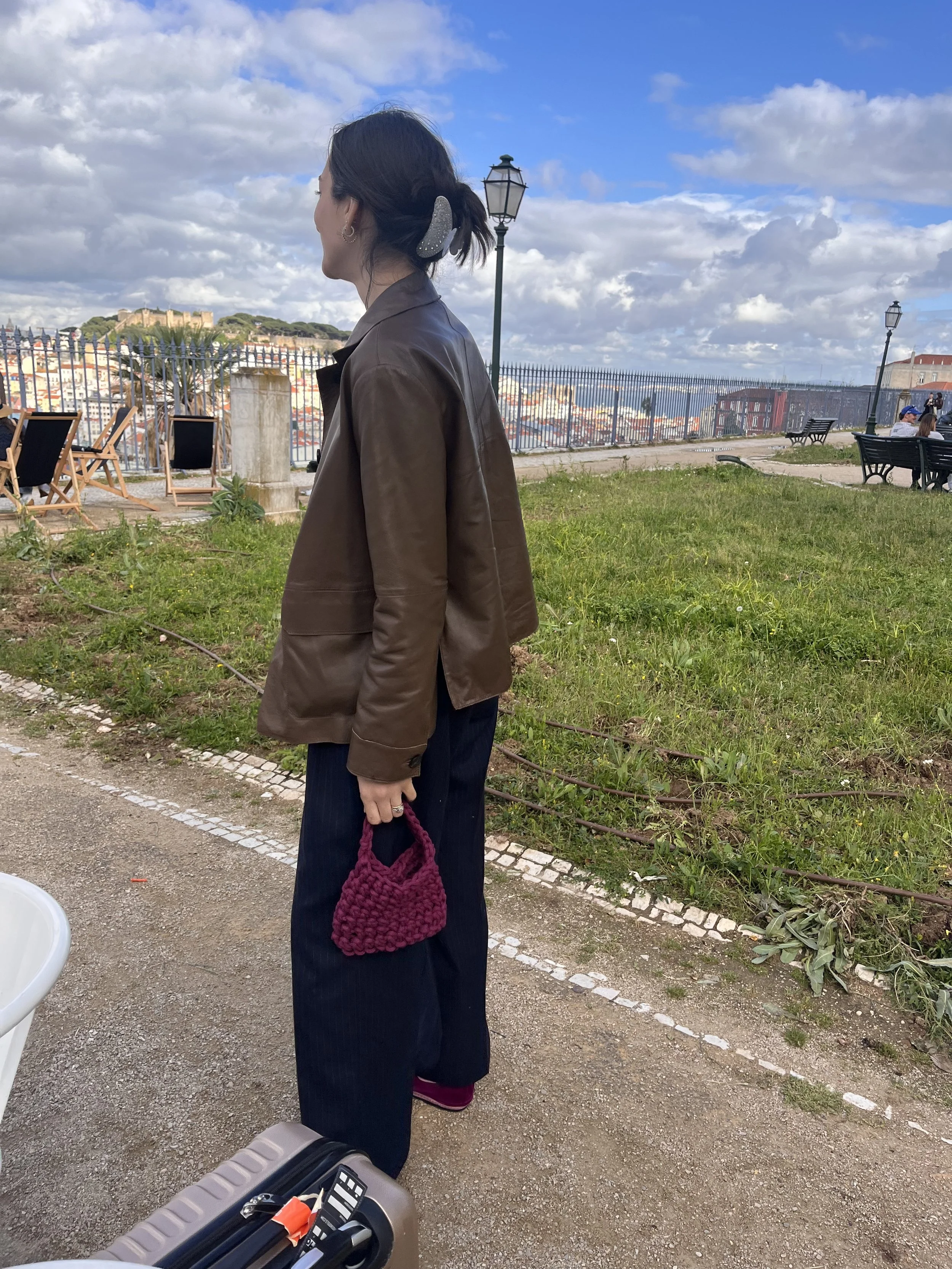 Eine Frau steht an einer Promenade mit Blick auf die Stadt, trägt eine braune Jacke, dunkle Hosen, hält eine rosa Stricktasche, und hat ihre Haare in einem Dutt mit Haarspange. Im Hintergrund sind Bänke, Straßenlaternen, eine Mauer und die Skyline si