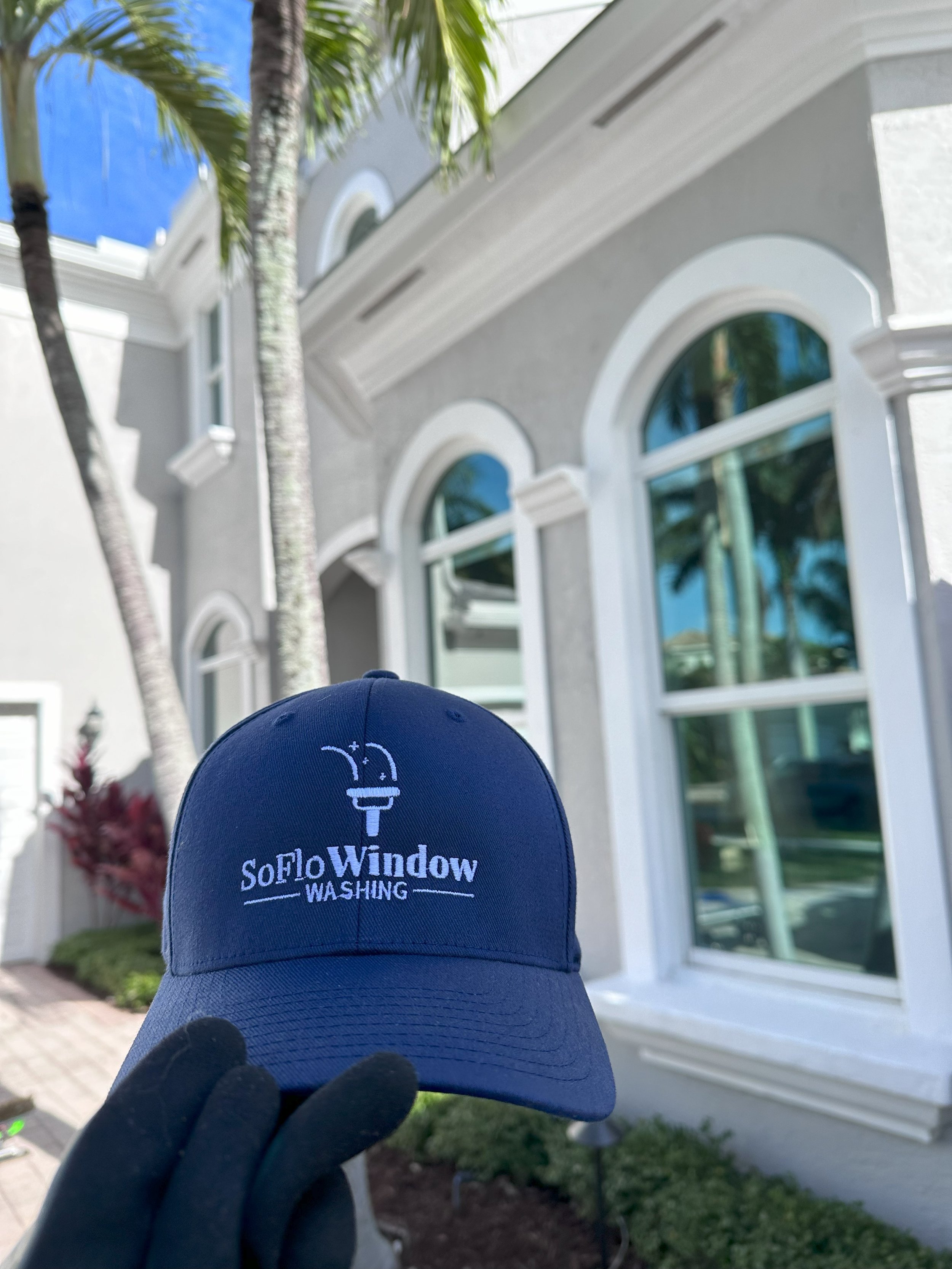 Person holding a SoFlo Window Washing cap in front of a house with arched windows and palm trees.