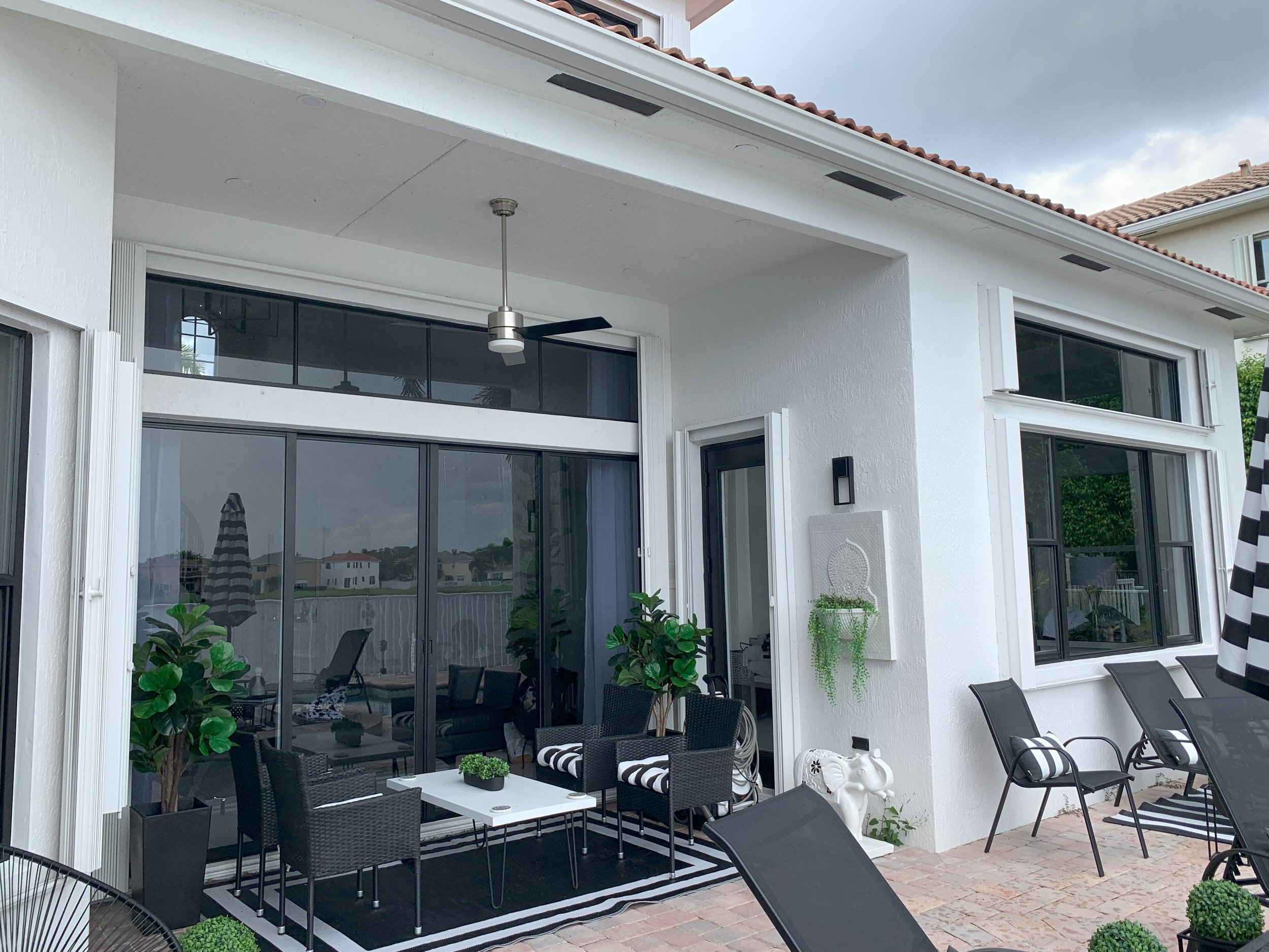 Outdoor patio with white walls, black wicker chairs, and a table. A ceiling fan is visible above, with potted plants and striped decor. Sliding glass doors lead inside the house. Brick flooring and a few lounge chairs complete the setting.