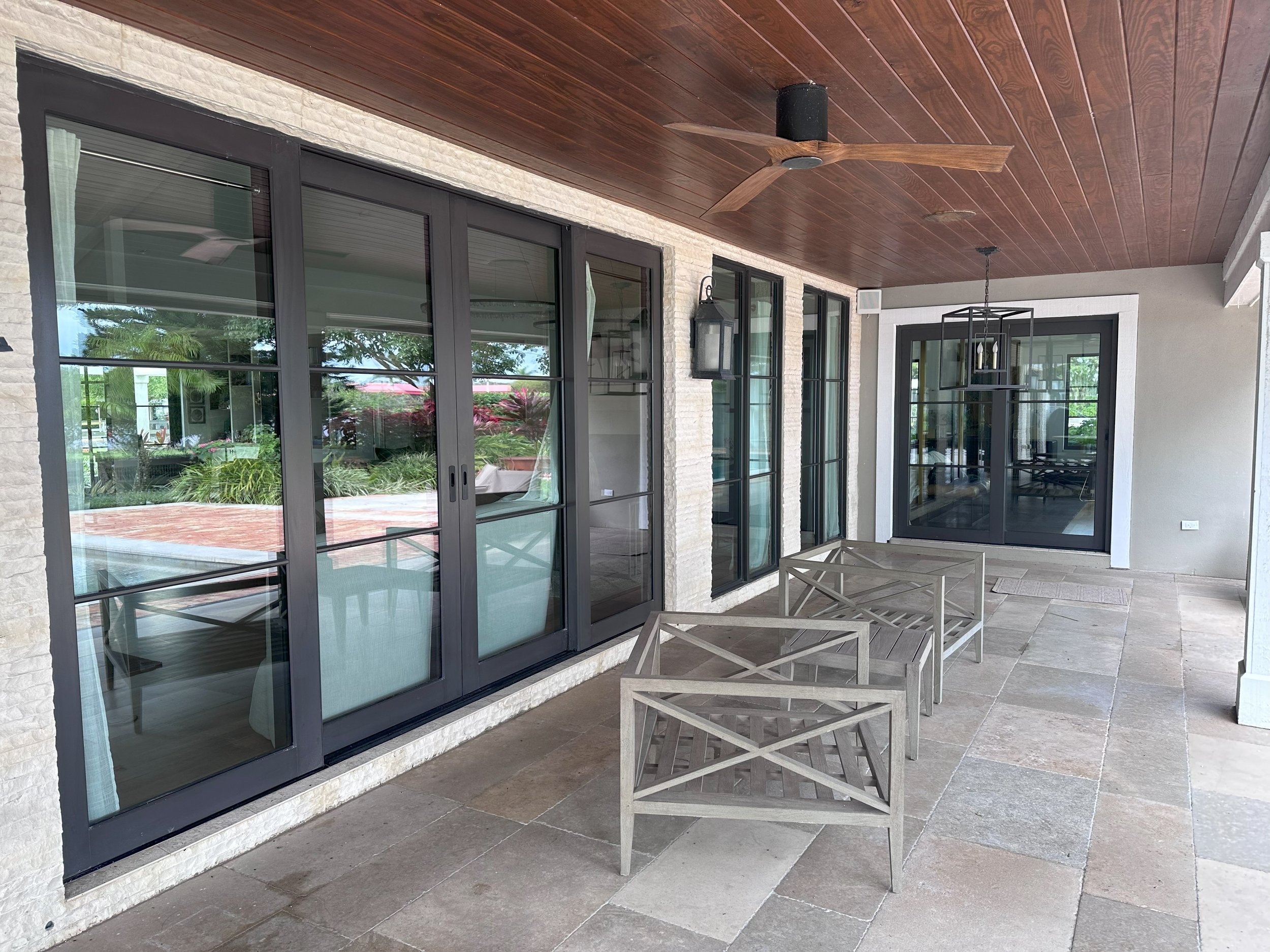 Modern patio with large glass doors, metal patio furniture, ceiling fan, and tiled flooring under a wooden-paneled ceiling.