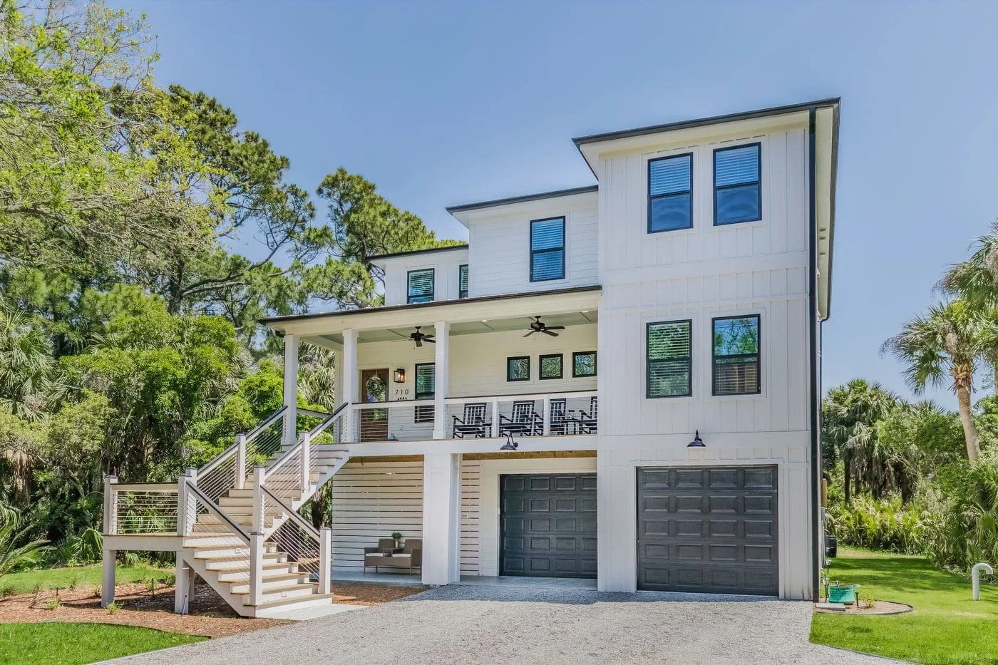 Two-story white modern house with elevated front porch, black garage doors, and surrounded by trees.