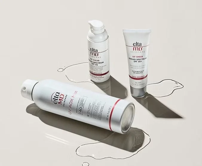 EltaMD skincare products including UV Clear, UV Sheer, and UV AOX Mist sunscreen bottles on a light background with liquid spills.