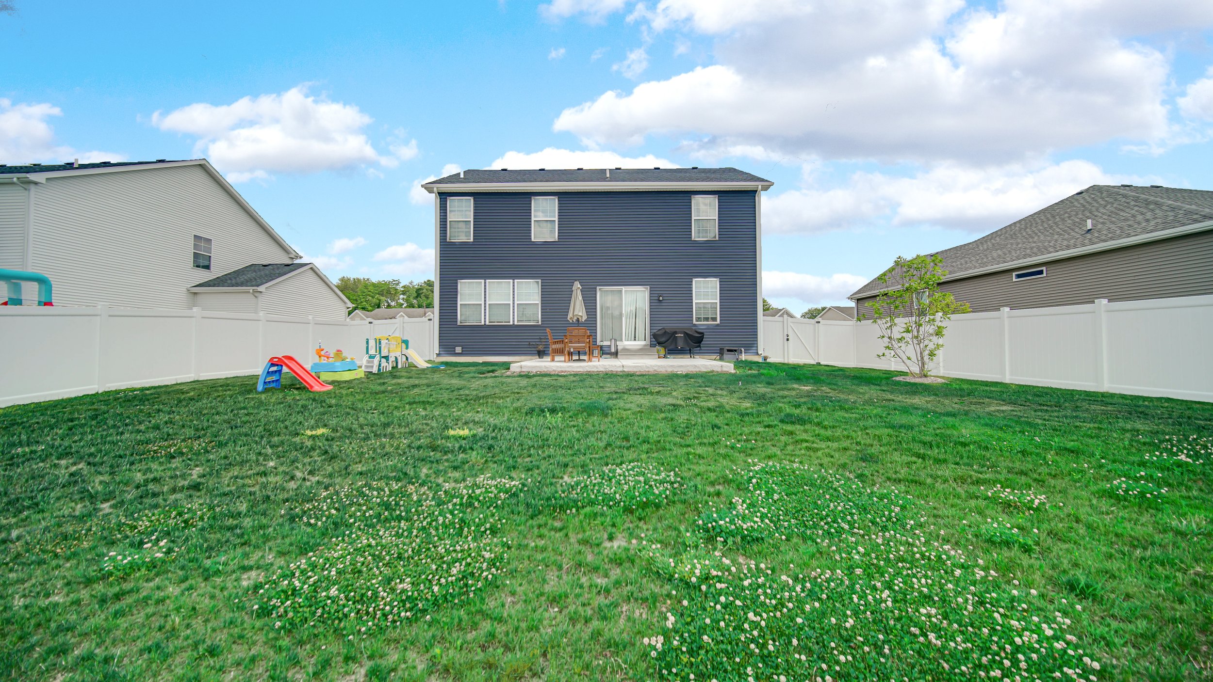 Backyard with a blue house, white fence, playset, table with umbrella, and outdoor furniture under a partly cloudy sky.