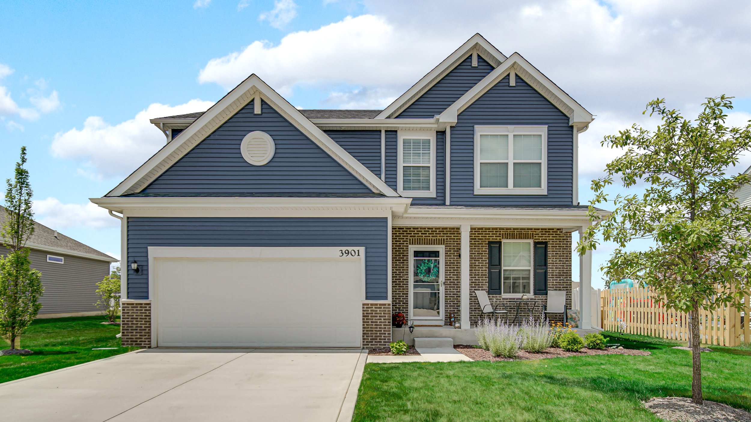 A two-story suburban house with blue siding, brick accents, white trim, and a front porch with outdoor chairs and a garden, under a partly cloudy sky.