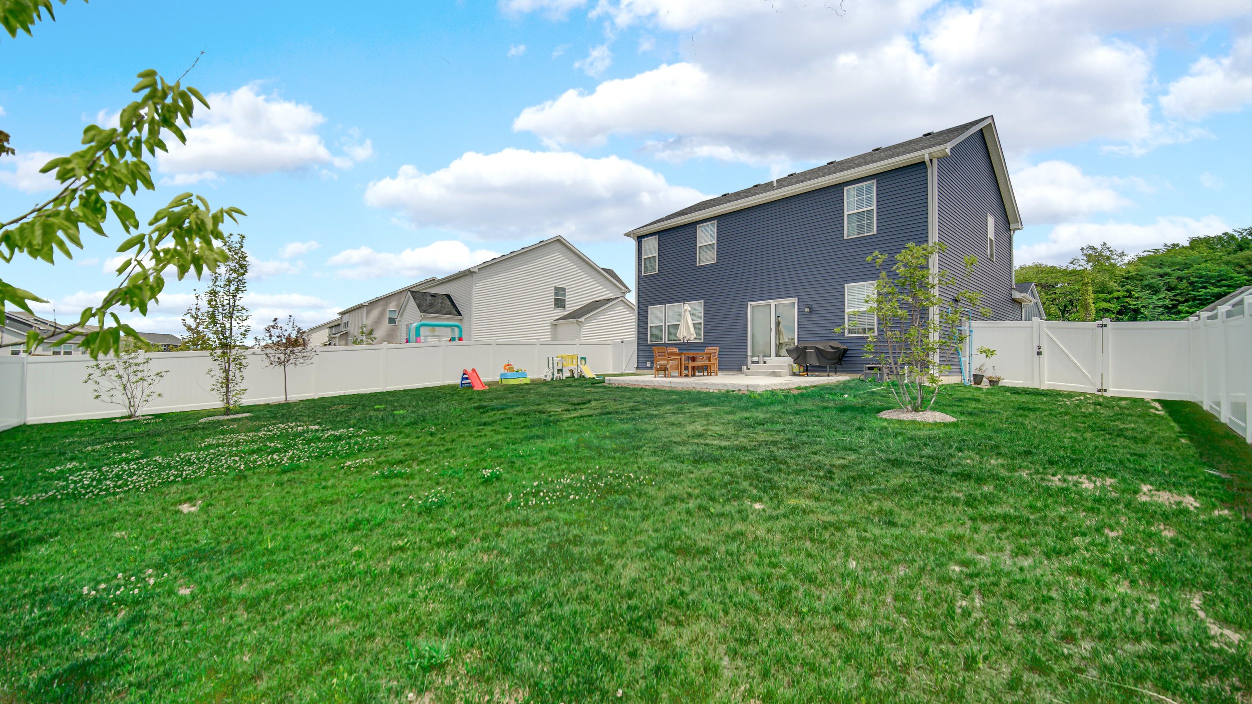 Backyard with green grass, small trees, a patio with outdoor furniture, children's play area, and neighboring houses under a partly cloudy sky.