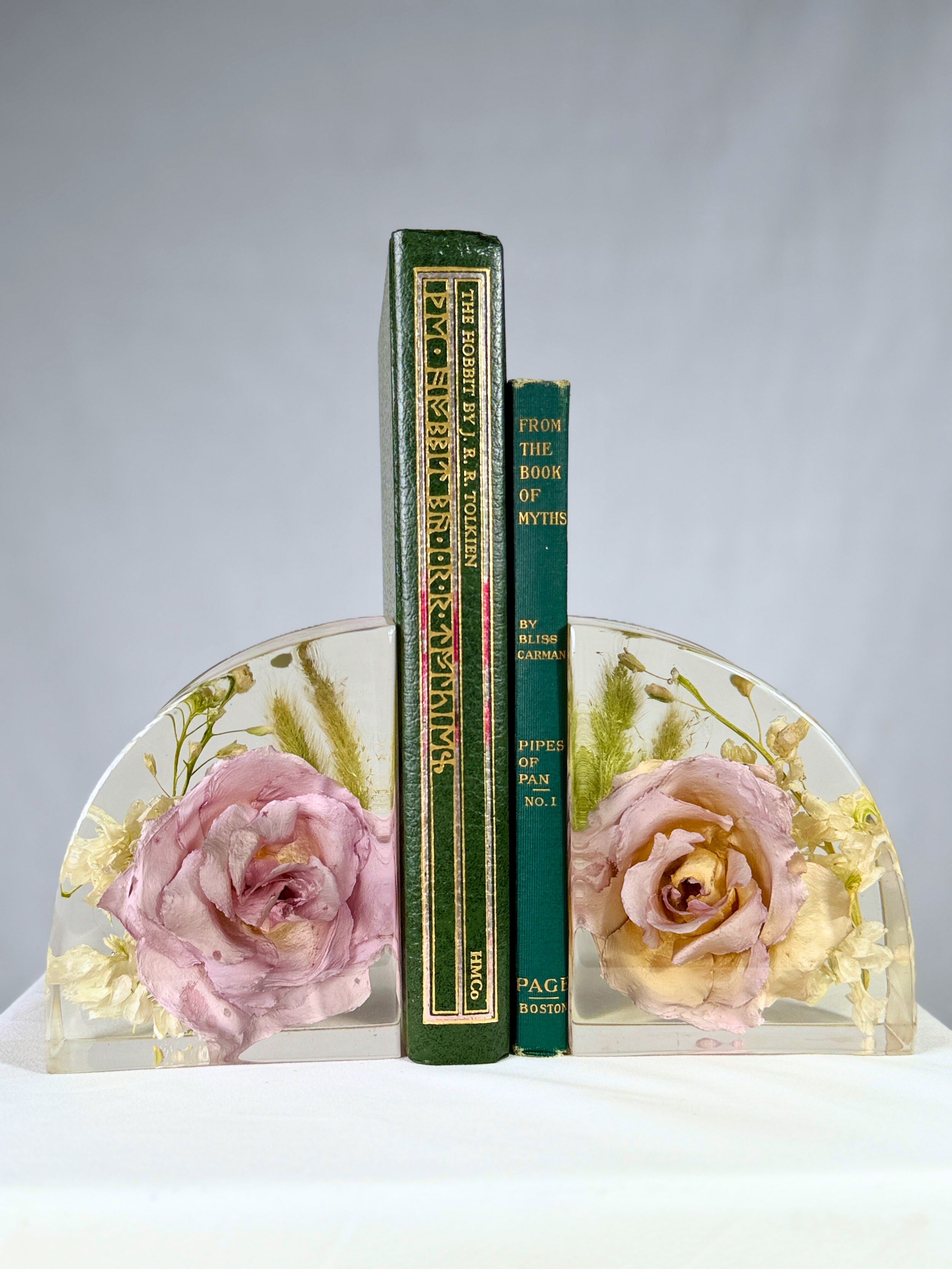 Three books displayed on a white surface, with two clear bookends containing pink and cream-colored dried roses and greenery.