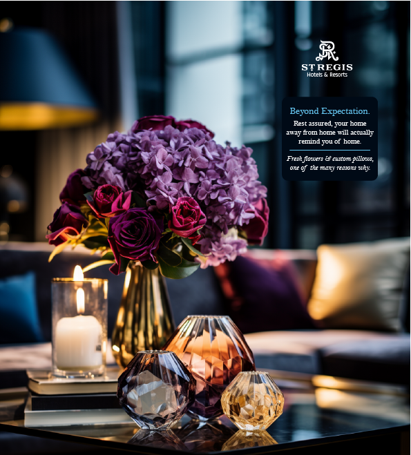 A floral arrangement with purple and pink flowers in a gold vase, surrounded by three colorful glass candle holders, on a black glass table in a modern, well-lit hotel lobby. A neon sign with the St. Regis Hotels & Resorts logo is visible in the background.