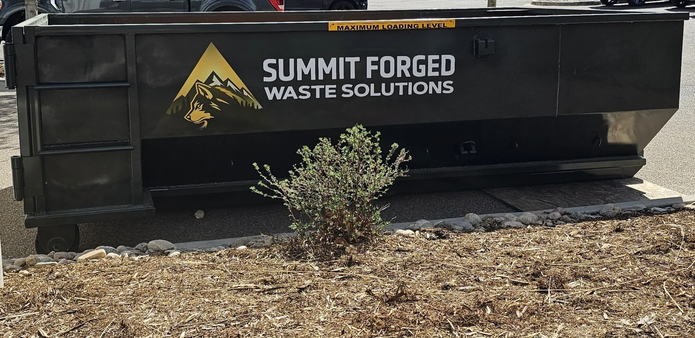 dumpster with white and yellow signage reading "SUMMIT FORGED WASTE SOLUTIONS" and a mountain and wolf logo. The dumpster is located outdoors, with a small shrub and soil in the foreground, and has a yellow "MAXIMUM LOADING LEVEL" label near the top.