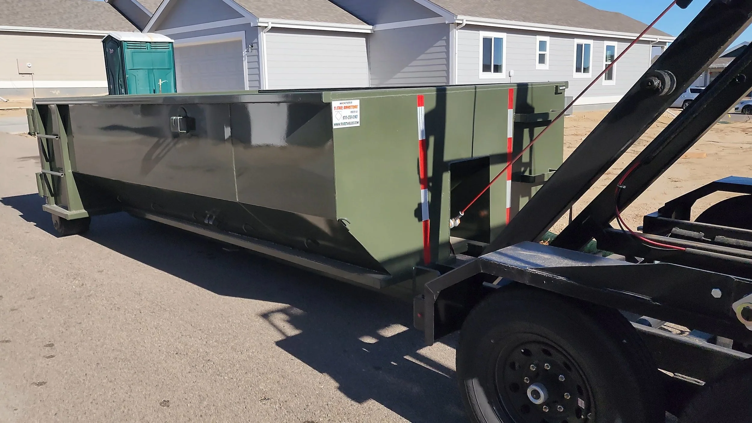 A large military-green dumpster with reflective red and white tape is mounted on a flatbed trailer at a construction site, with a partially built residential neighborhood in the background.