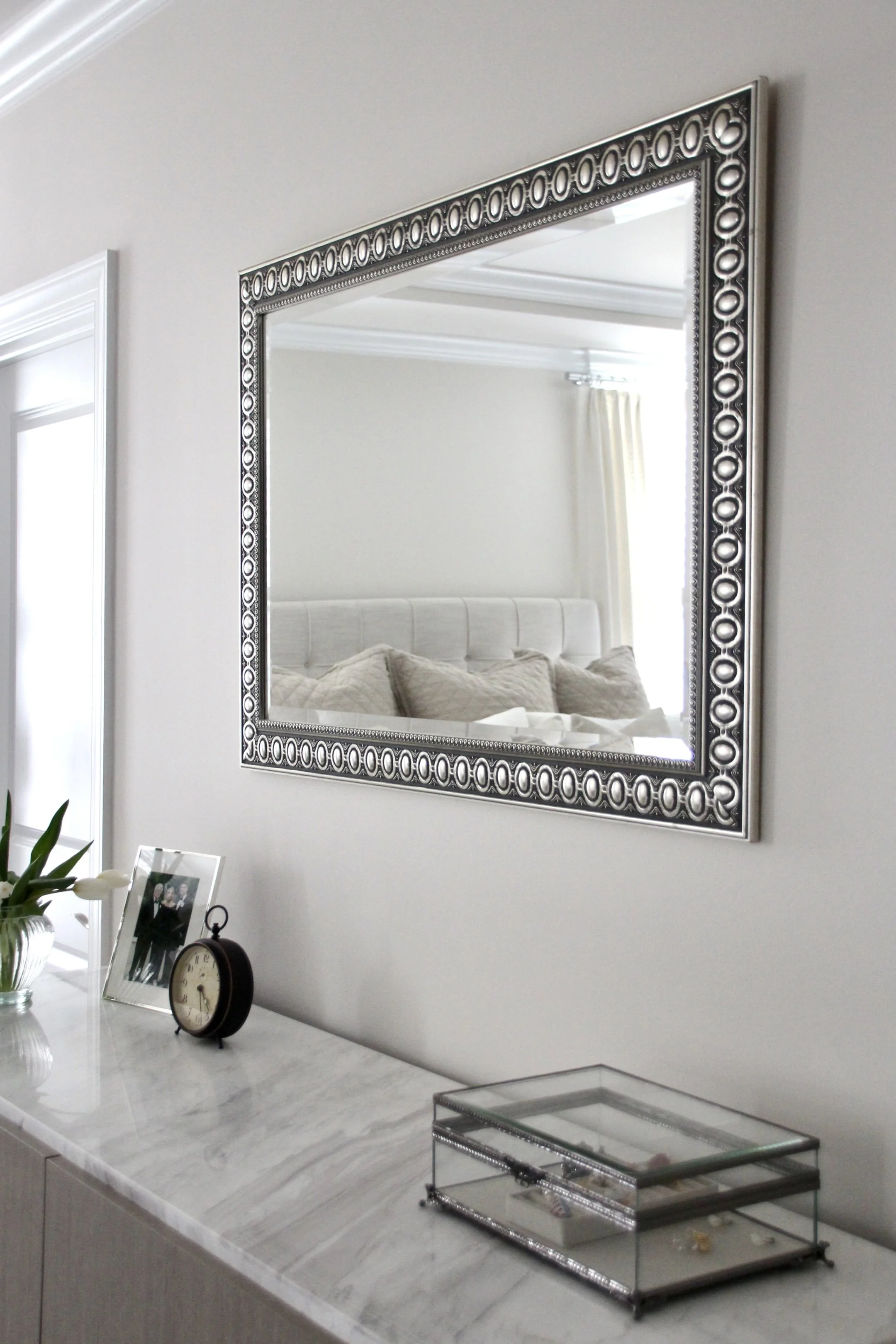 Decorative mirror with ornate frame above a marble surface with an antique-style clock, a glass box, and a vase with white flowers.