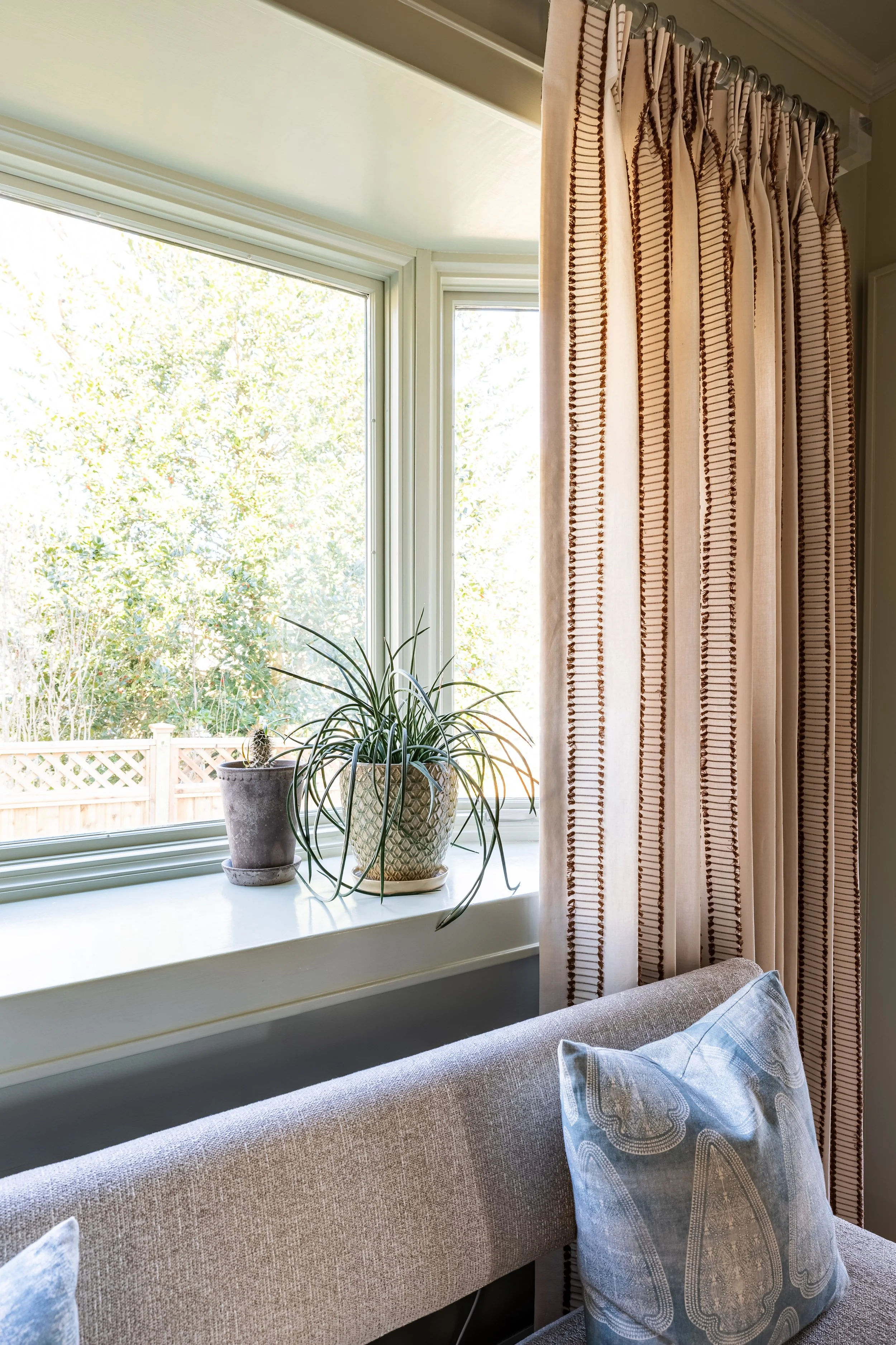 A cozy interior showing a patterned curtain next to a window. On the window sill, there are two potted plants, including one with long, thin leaves. A gray upholstered bench with a blue decorative pillow is partially visible below the window.