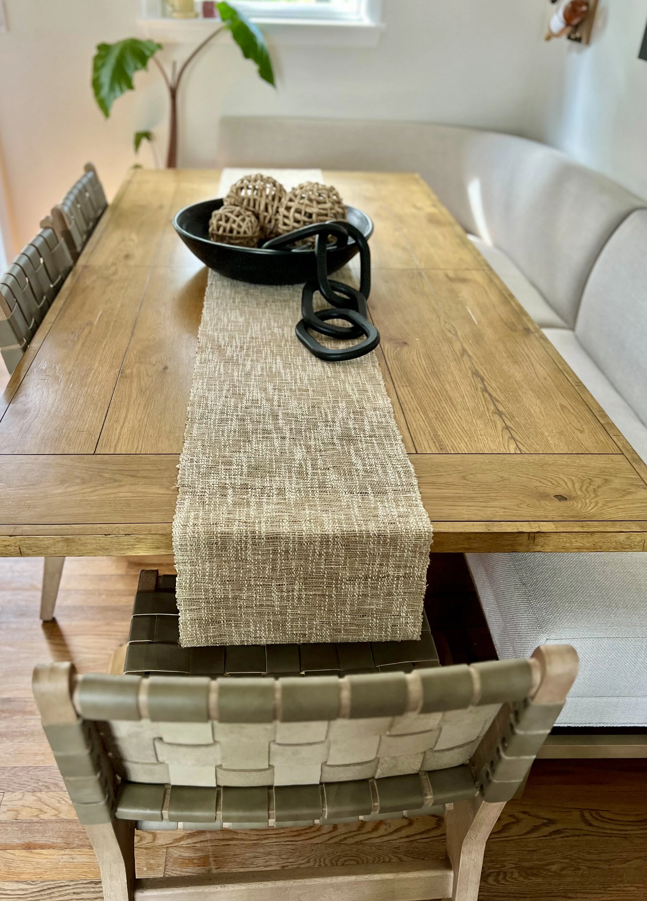 Dining table setup with wooden table, beige table runner, woven chairs, black bowl with decorative wicker orbs, and a black decorative chain. A potted plant is visible in the background.