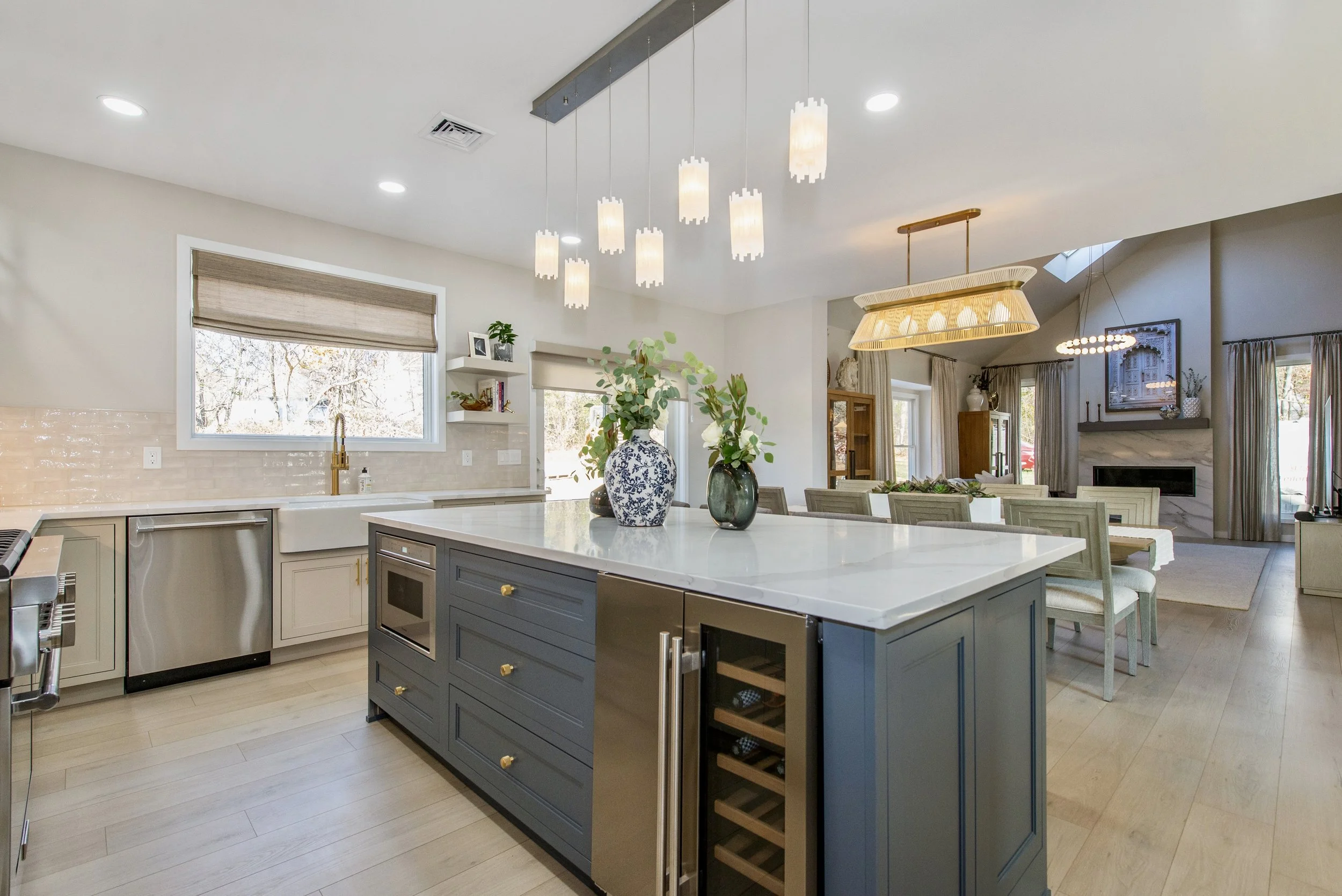 Modern kitchen with a large island, pendant lights, built-in appliances, and decorative vases. Adjacent dining area with a wooden table, chairs, and a fireplace in the background.